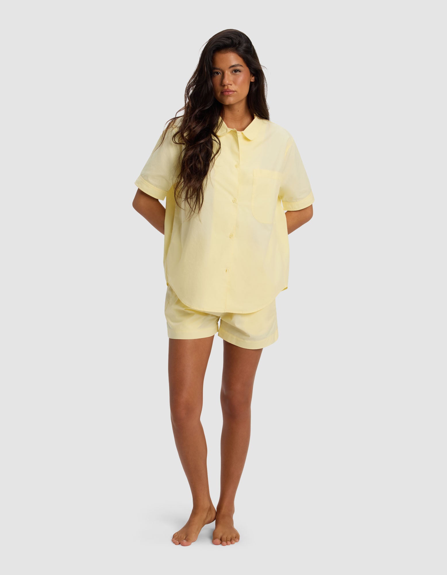 A woman stands barefoot against a plain background, wearing the Cozy Earth Women's Sutton Short Sleeve Pajama Set in light yellow, with her hands in her pockets.