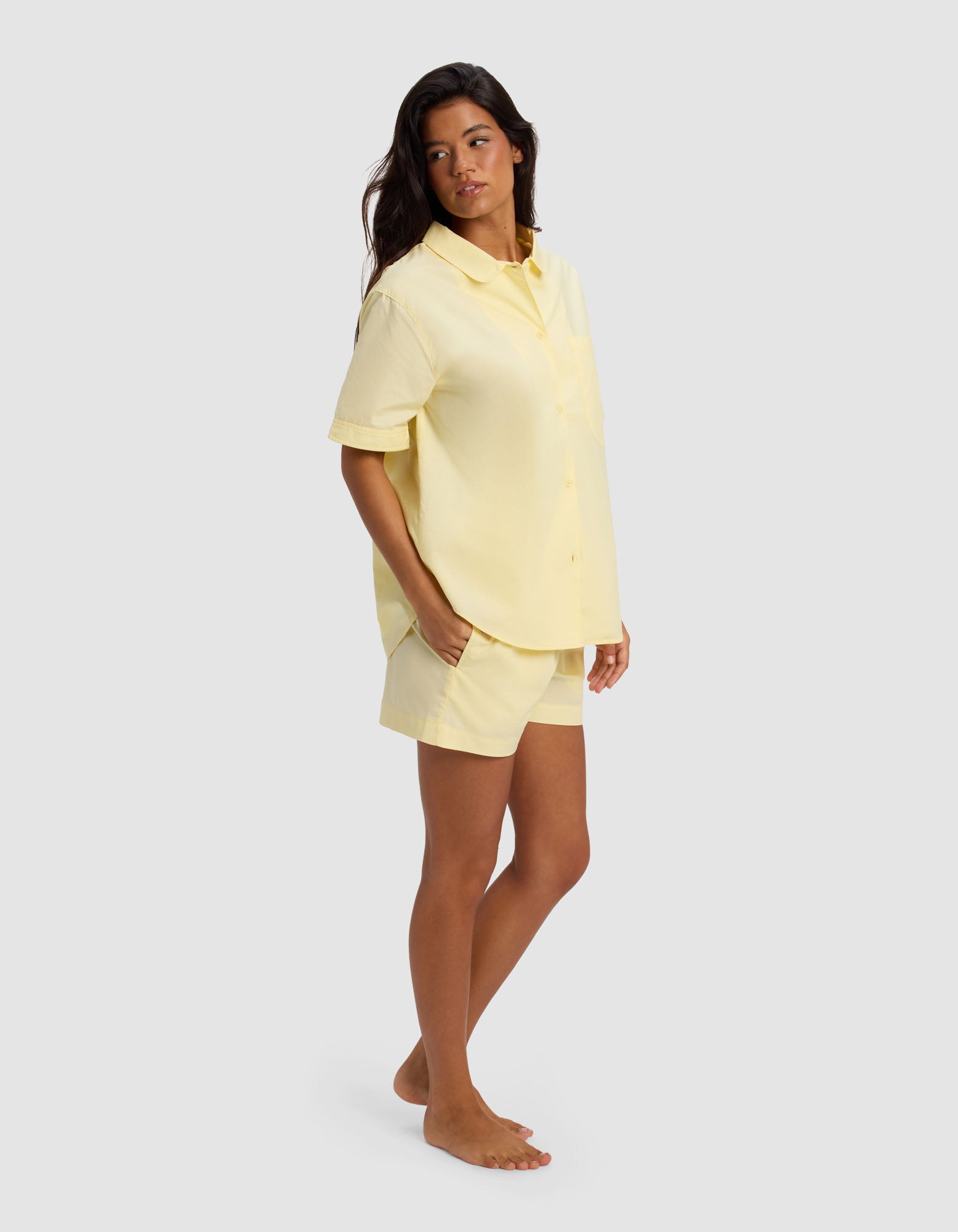 A woman stands barefoot against a light gray background, wearing the Cozy Earth Women's Sutton Short Sleeve Pajama Set in yellow, with her hands in her pockets and looking slightly to the side.