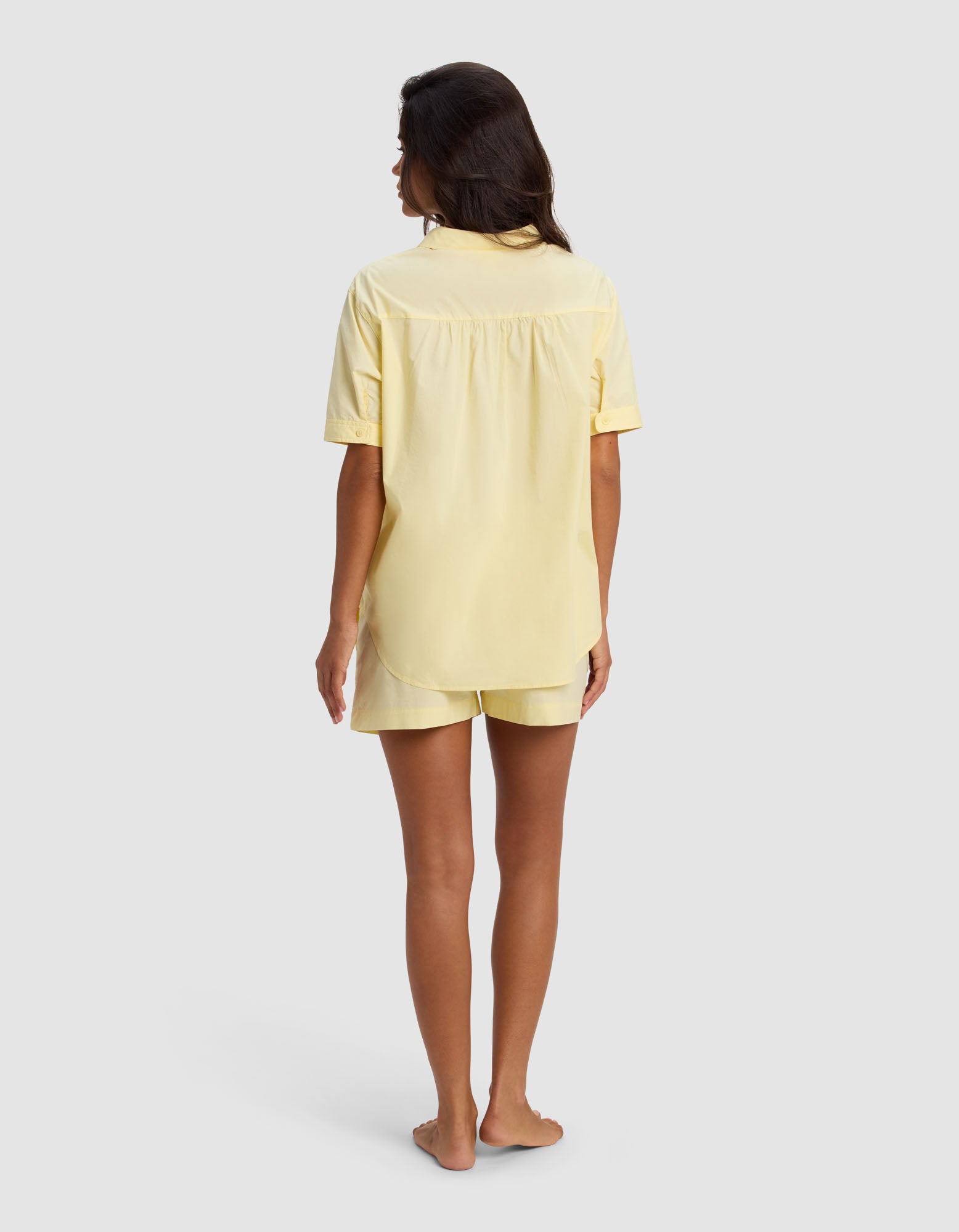 A woman with long dark hair stands barefoot, facing away, wearing the Cozy Earth Women's Sutton Short Sleeve Pajama Set in light yellow against a plain light gray background.