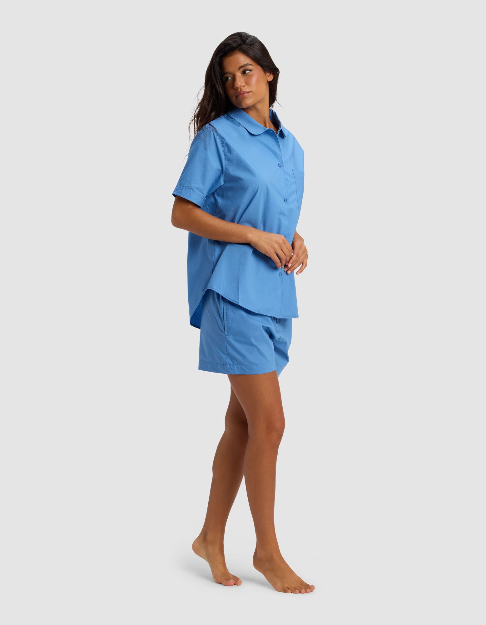 A woman stands barefoot on a light gray background, looking to her left, wearing Cozy Earth's Women's Sutton Short Sleeve Pajama Set—a loose-fitting blue button-up shirt with matching blue shorts.