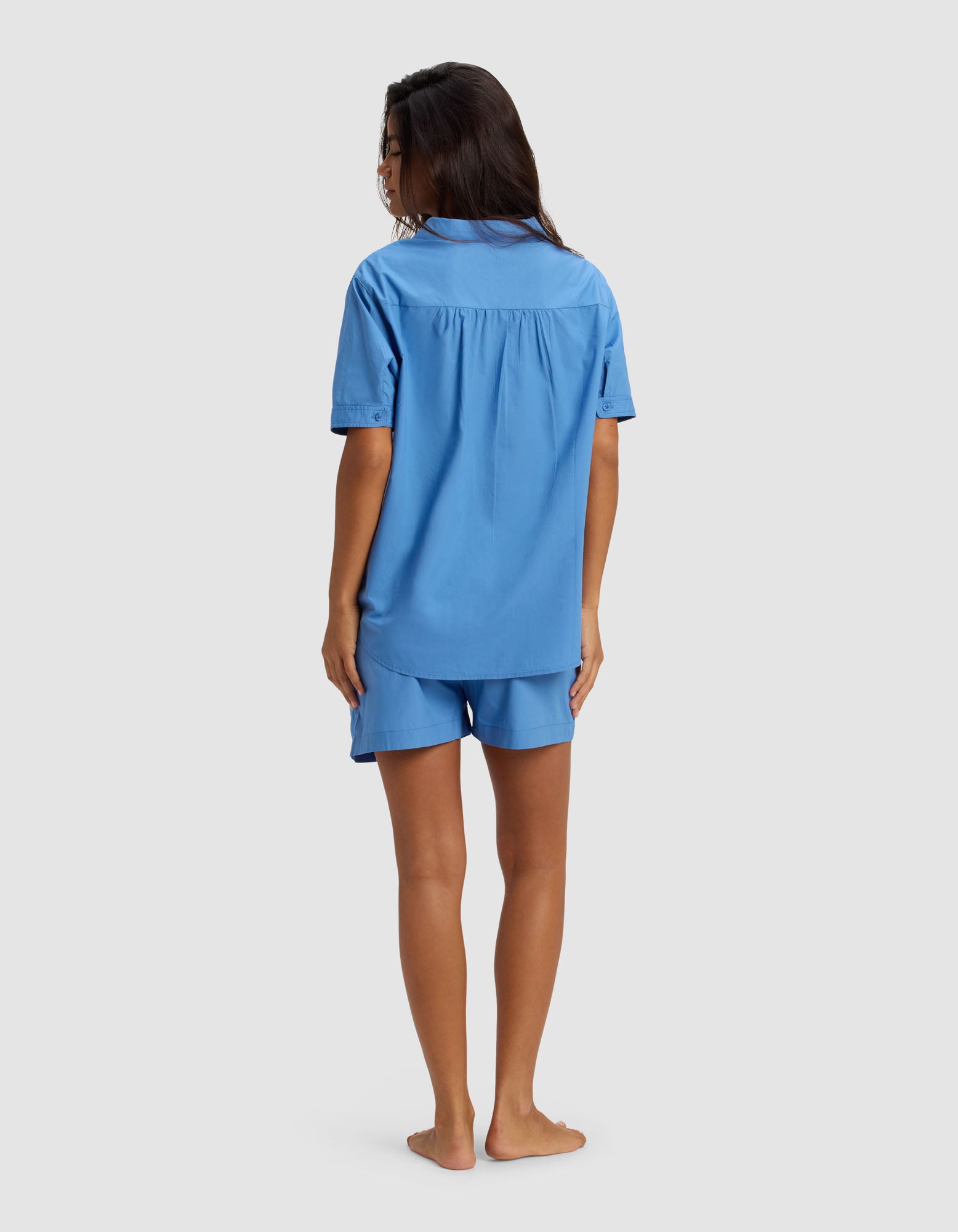 A woman stands barefoot, facing away, wearing the Cozy Earth Women's Sutton Short Sleeve Pajama Set in light blue, featuring a loose-fit shirt and matching shorts against a plain light gray background.