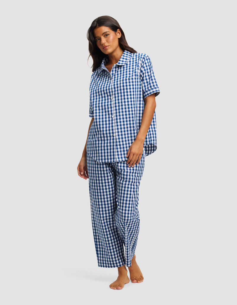 A woman stands barefoot against a light gray background, wearing the Cozy Earth Women's Sutton Short Sleeve & Pant Pajama Set in a blue and white checkered pattern.