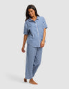 A woman with long dark hair smiles while looking down, barefoot in the Cozy Earth Women's Sutton Short Sleeve & Pant Pajama Set—a blue and white checkered collared button-up shirt and loose pants.|Color:Pacific Gingham