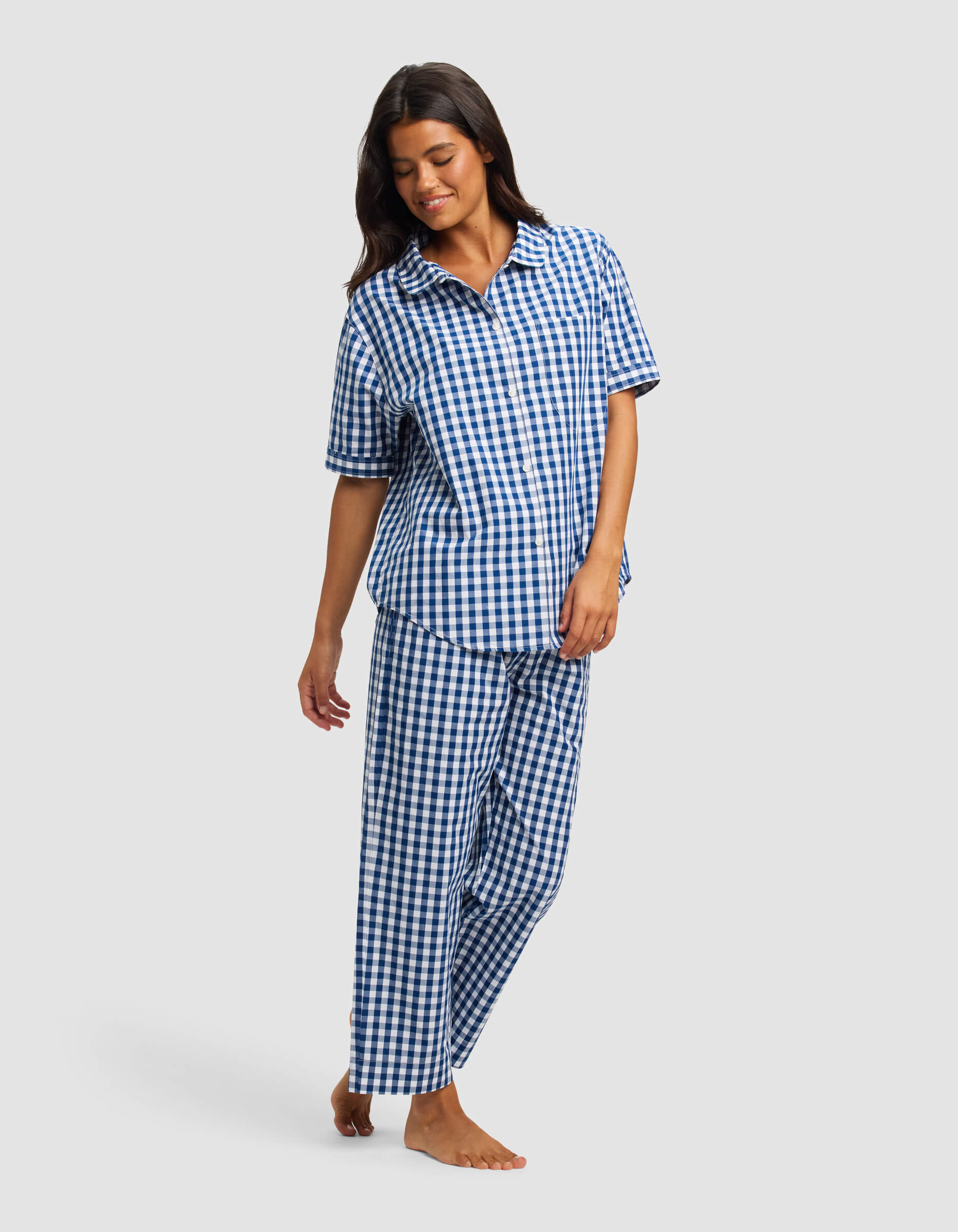 A woman models the Cozy Earth Women's Sutton Short Sleeve & Pant Pajama Set, featuring a blue and white checkered design, standing barefoot against a plain light gray background.|Color:Pacific Gingham