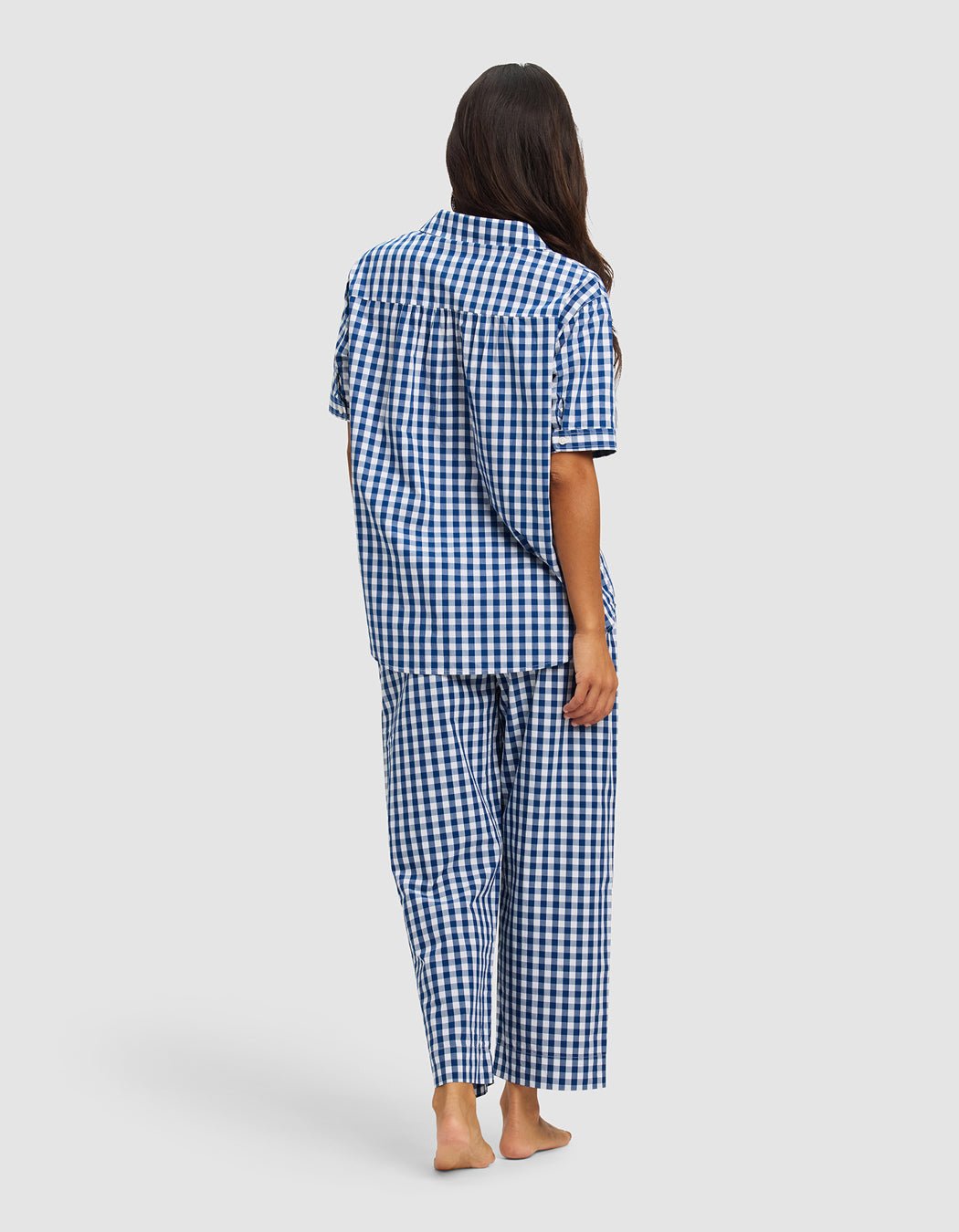 A woman with long brown hair stands barefoot, facing away, wearing Cozy Earth’s Women's Sutton Short Sleeve & Pant Pajama Set in blue and white checks against a plain light background.