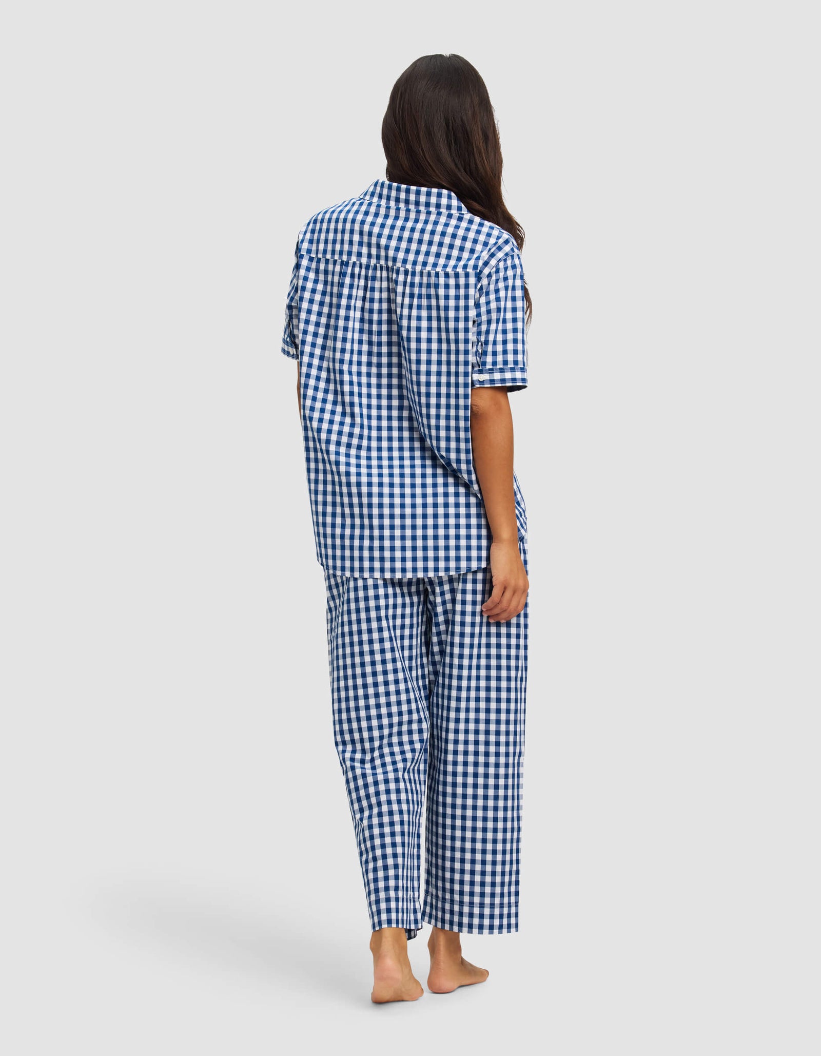 A woman with long dark hair stands barefoot, facing away, wearing the Cozy Earth Women's Sutton Short Sleeve & Pant Pajama Set in blue and white checks, against a plain light background.