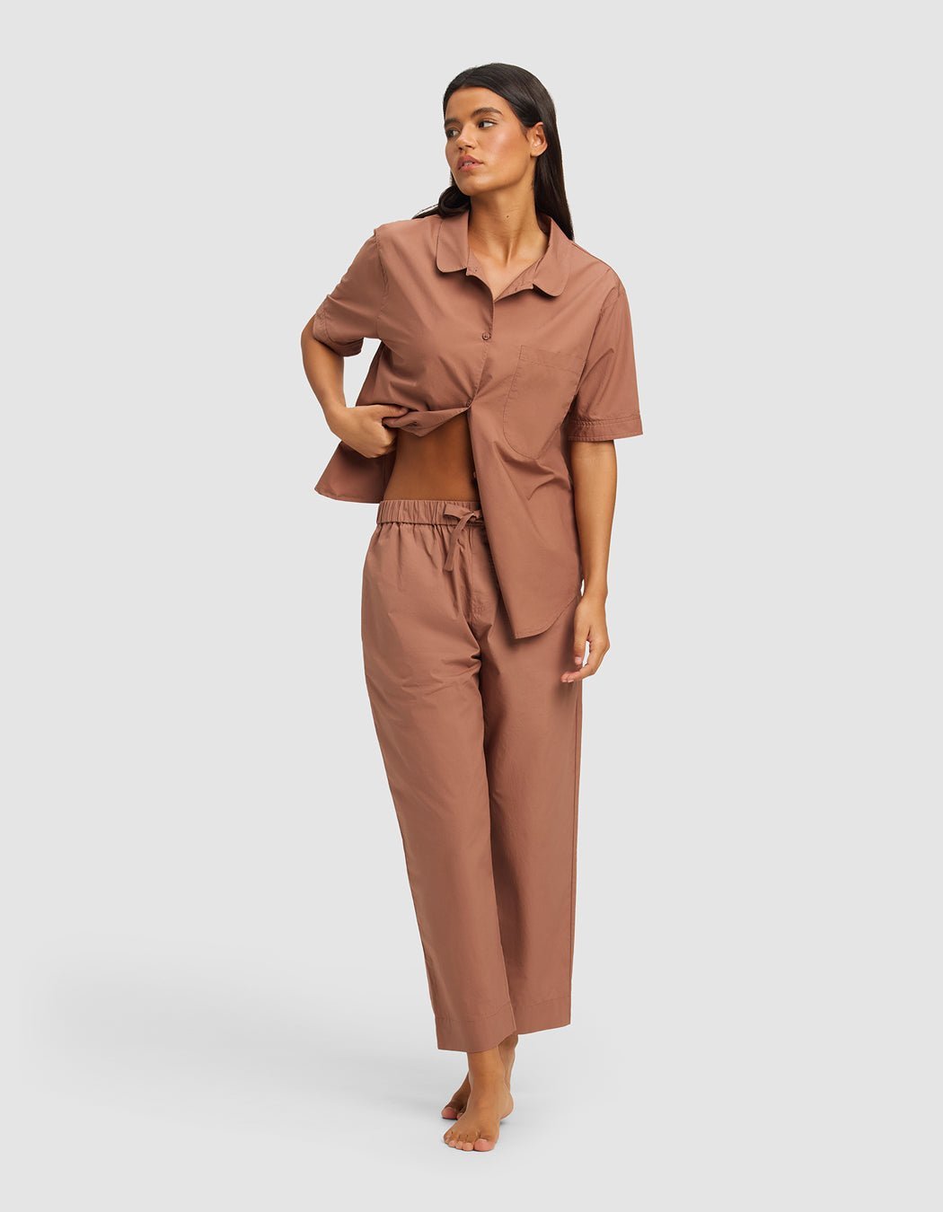 A woman, barefoot and looking to the side, wears the Cozy Earth Women's Sutton Short Sleeve & Pant Pajama Set—featuring a loose fit, collared top, and elastic waist pants—while lifting part of the shirt.