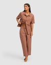 A woman, barefoot and looking to the side, wears the Cozy Earth Women's Sutton Short Sleeve & Pant Pajama Set—featuring a loose fit, collared top, and elastic waist pants—while lifting part of the shirt.|Color:Saddle