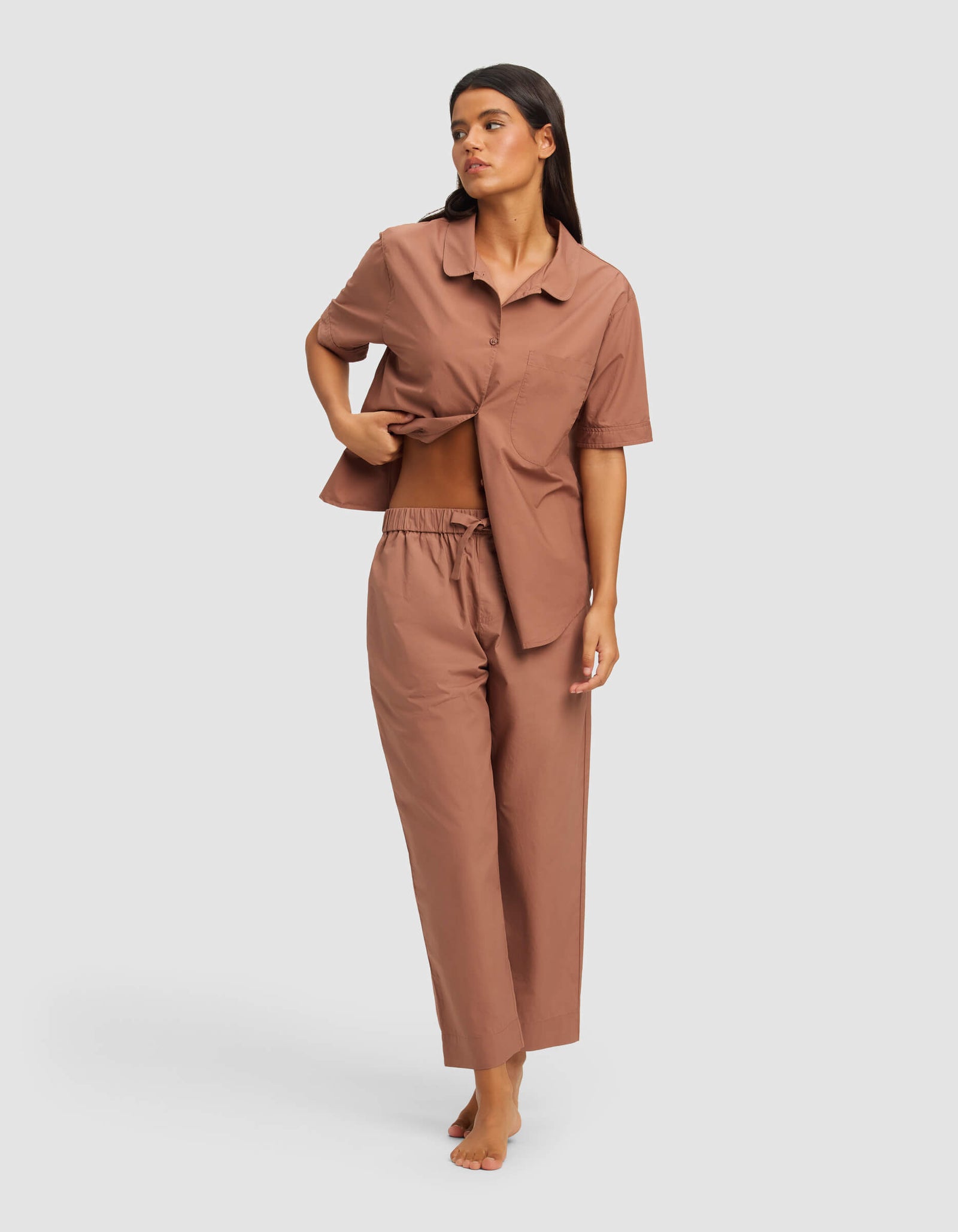 A woman stands barefoot against a plain background wearing the Cozy Earth Women's Sutton Short Sleeve & Pant Pajama Set in brown, lifting her shirt to reveal her midriff.