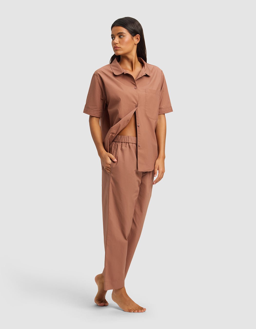 A woman stands barefoot in the Cozy Earth Women's Sutton Short Sleeve & Pant Pajama Set, a loose-fitting brown button-up shirt with matching pants. She looks to the side with one hand in her pocket and the other gently holding her shirt open. |Color:Saddle