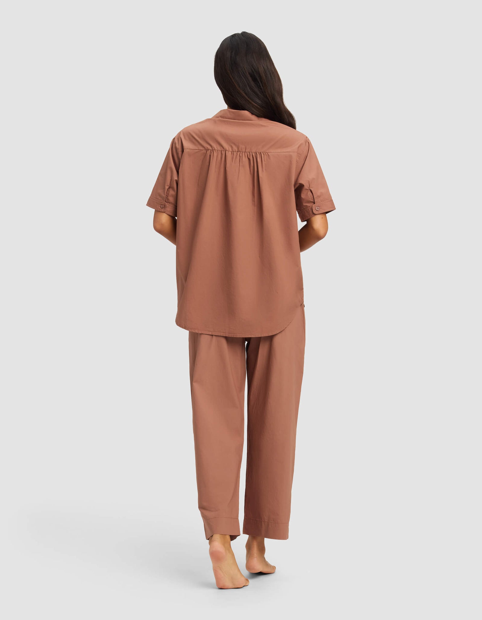 A woman with long dark hair stands barefoot, facing away from the camera, wearing the Cozy Earth Women's Sutton Short Sleeve & Pant Pajama Set against a plain light background.