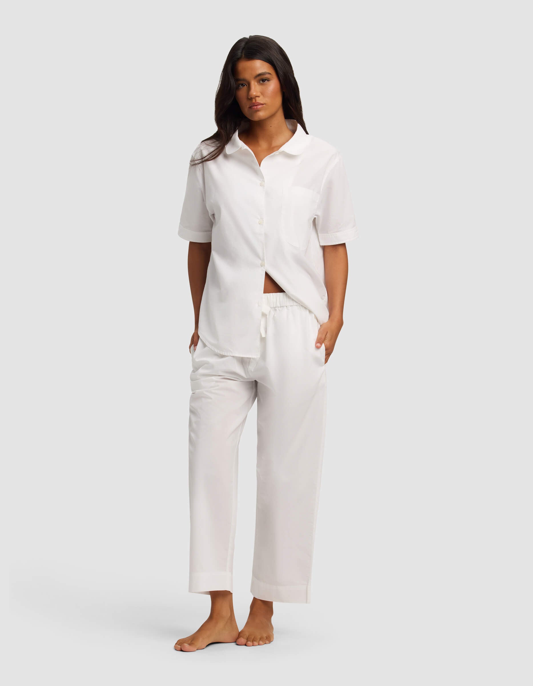 A woman stands barefoot against a plain background, wearing the Cozy Earth Women's Sutton Short Sleeve & Pant Pajama Set in white. The loose-fitting set features a button-up shirt, short sleeves, and matching drawstring pants. Her hands are in her pockets.