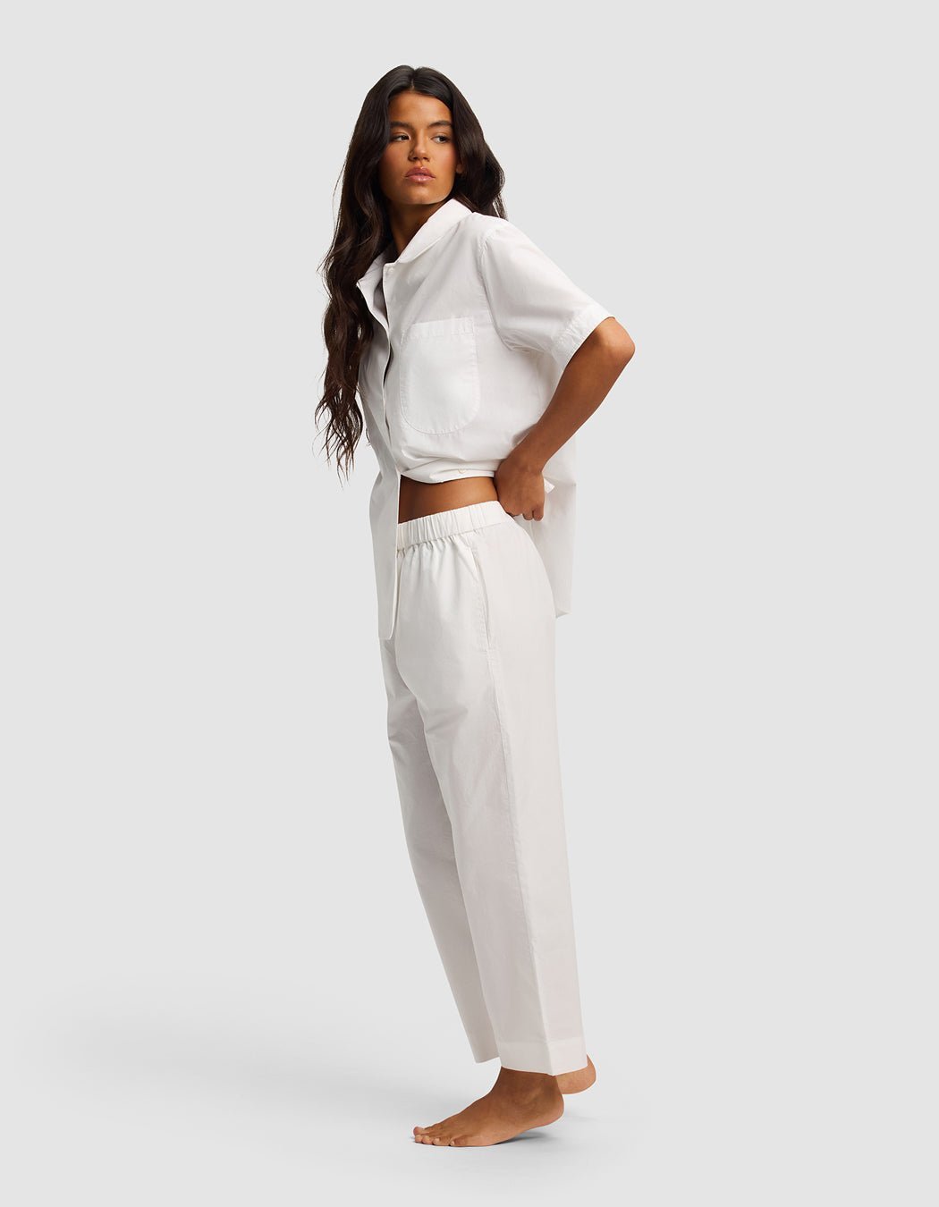 A woman with long, wavy brown hair stands barefoot against a plain background, wearing the Cozy Earth Women's Sutton Short Sleeve & Pant Pajama Set in white. She is turned slightly to the side and looks ahead.
