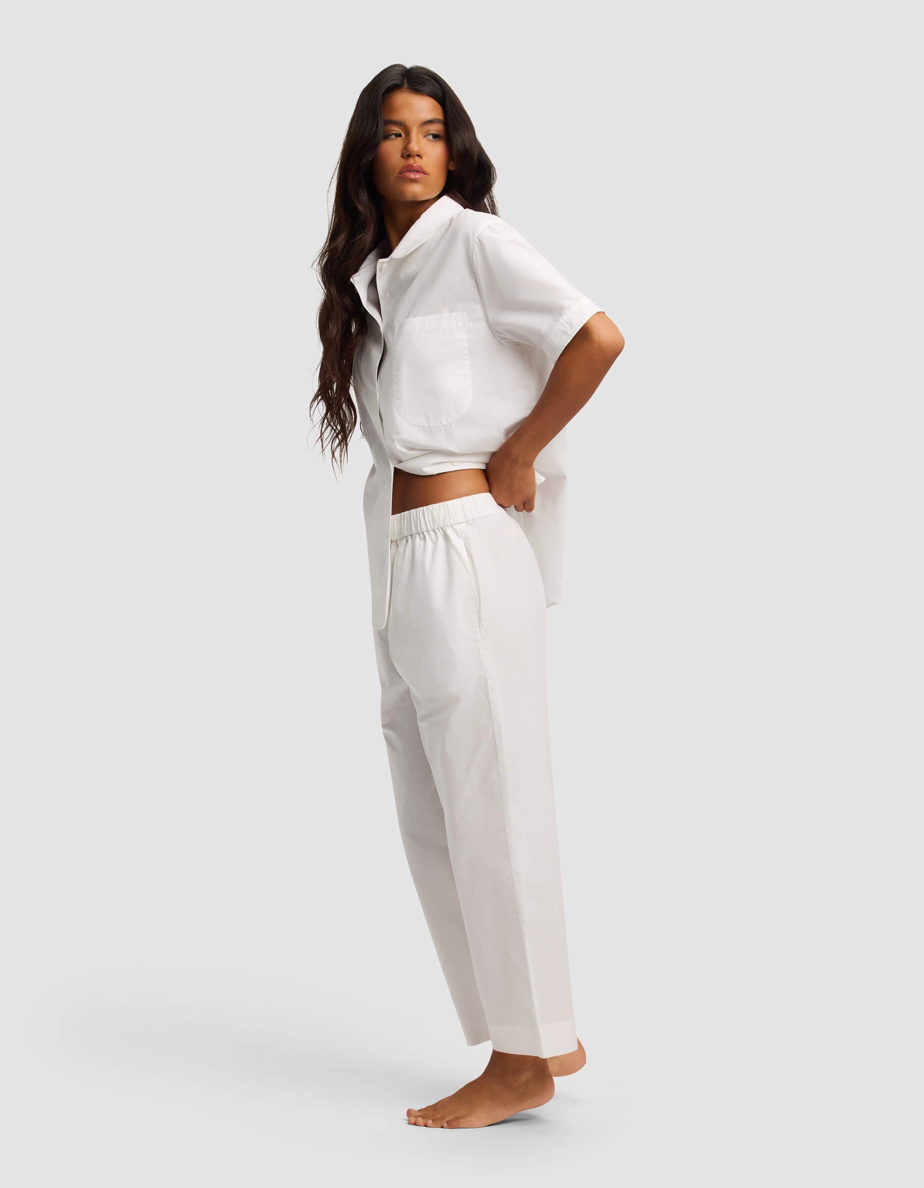 A woman with long dark hair stands barefoot, posing sideways against a plain light background in the Cozy Earth Women's Sutton Short Sleeve & Pant Pajama Set.|Color:White