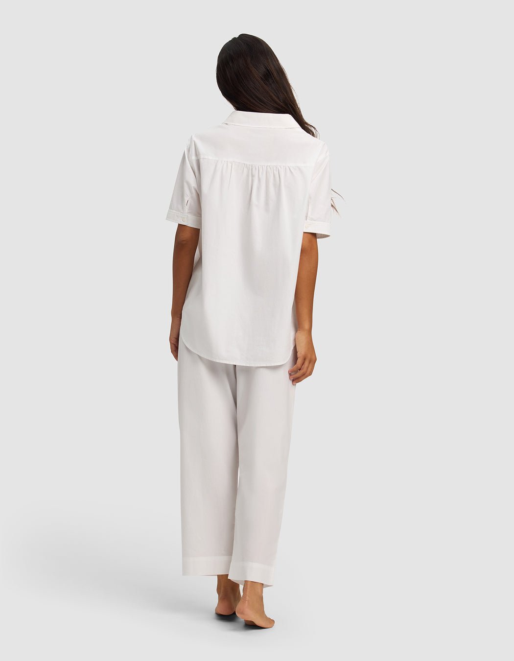 A person with long dark hair stands barefoot, facing away, wearing the Cozy Earth Women's Sutton Short Sleeve & Pant Pajama Set in white against a plain light gray background.