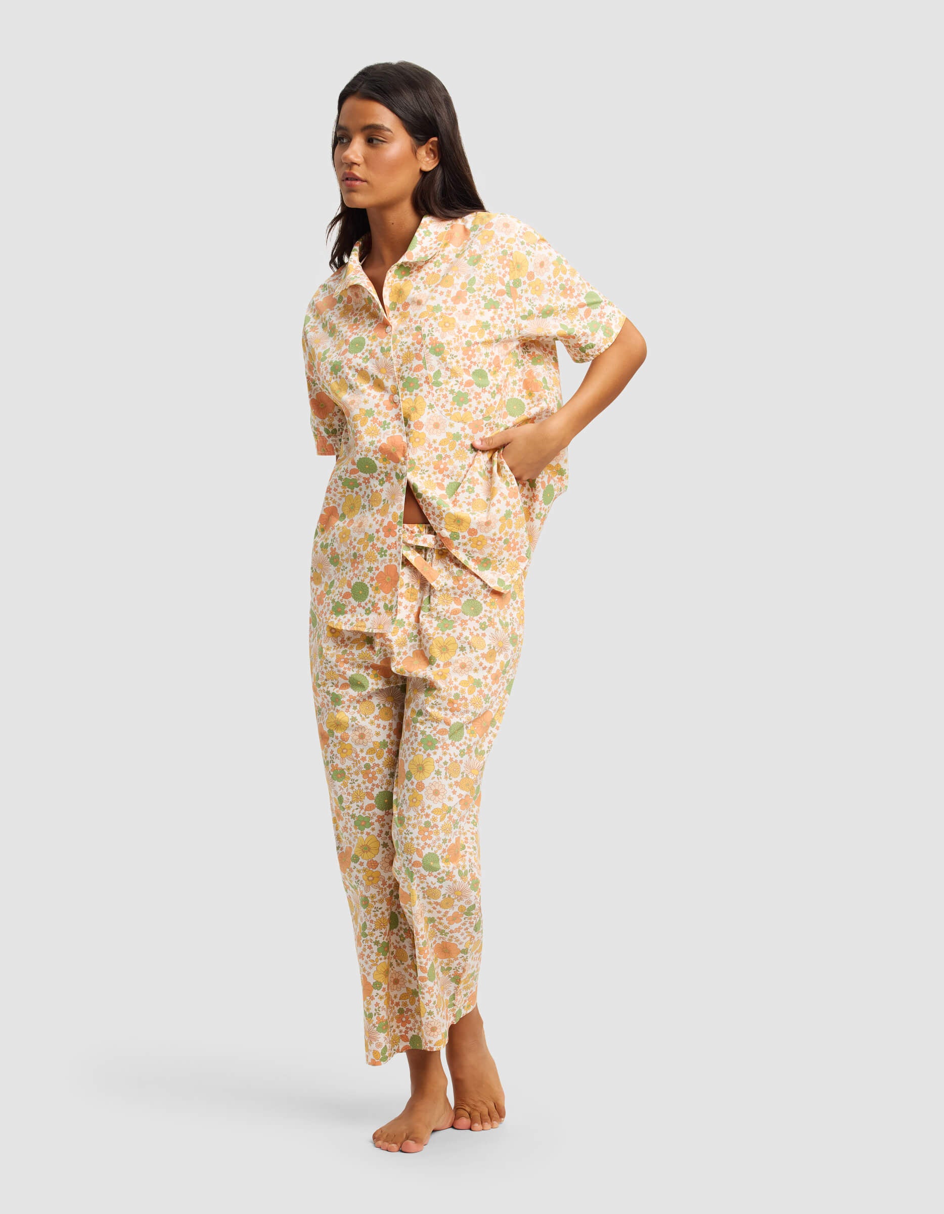 A woman stands barefoot in the Cozy Earth Women's Sutton Short Sleeve & Pant Pajama Set—matching floral pajamas with a pastel short-sleeve button-up top and pants—posing with one hand in her pocket against a plain light background.|Color:Wildflower