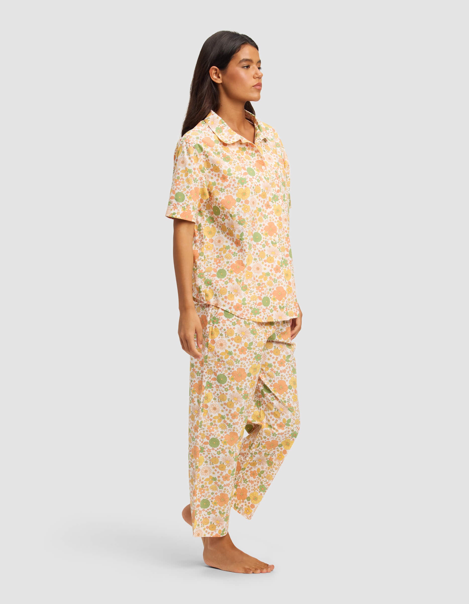 A woman is barefoot in a Cozy Earth Women's Sutton Short Sleeve & Pant Pajama Set, featuring a colorful floral pattern in peach, green, and white. The set includes a button-up short-sleeve shirt and loose pants, set against a plain light gray background.|Color:Wildflower