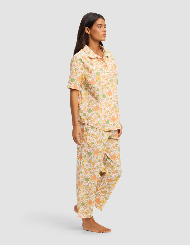 A woman is barefoot in a Cozy Earth Women's Sutton Short Sleeve & Pant Pajama Set, featuring a colorful floral pattern in peach, green, and white. The set includes a button-up short-sleeve shirt and loose pants, set against a plain light gray background.