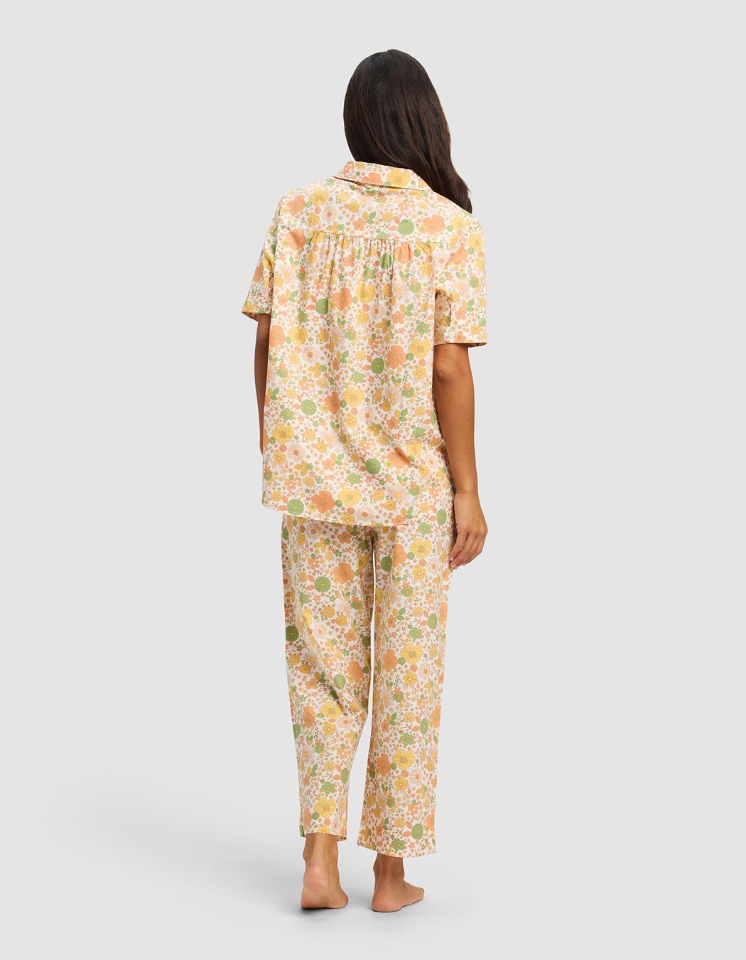 A woman with long brown hair stands barefoot, facing away, wearing Cozy Earth's Women's Sutton Short Sleeve & Pant Pajama Set in a soft orange, green, and white floral pattern on a light background.