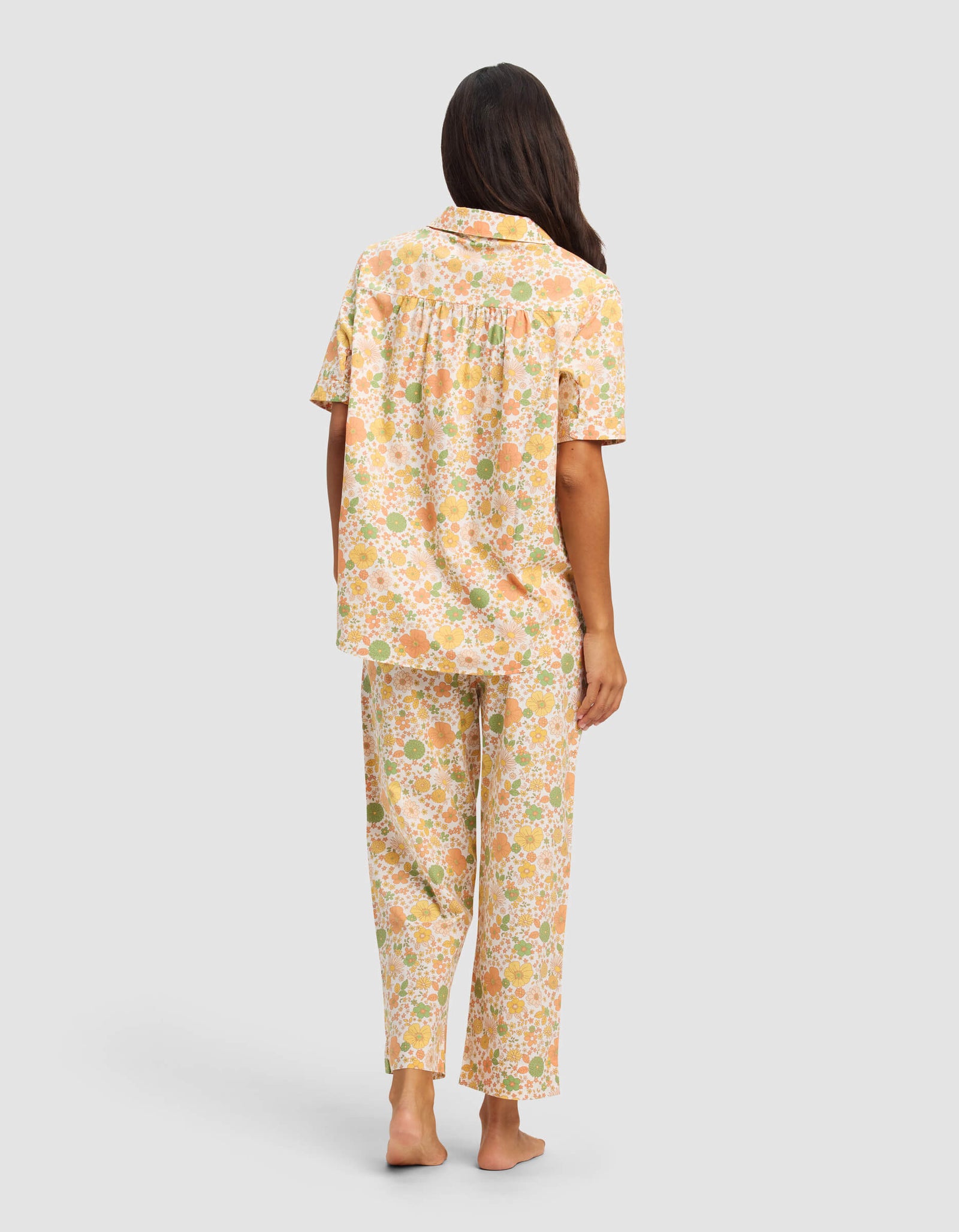A woman stands barefoot facing away, wearing the Cozy Earth Women's Sutton Short Sleeve & Pant Pajama Set in a pastel peach, green, and white floral print, set against a plain gray background.