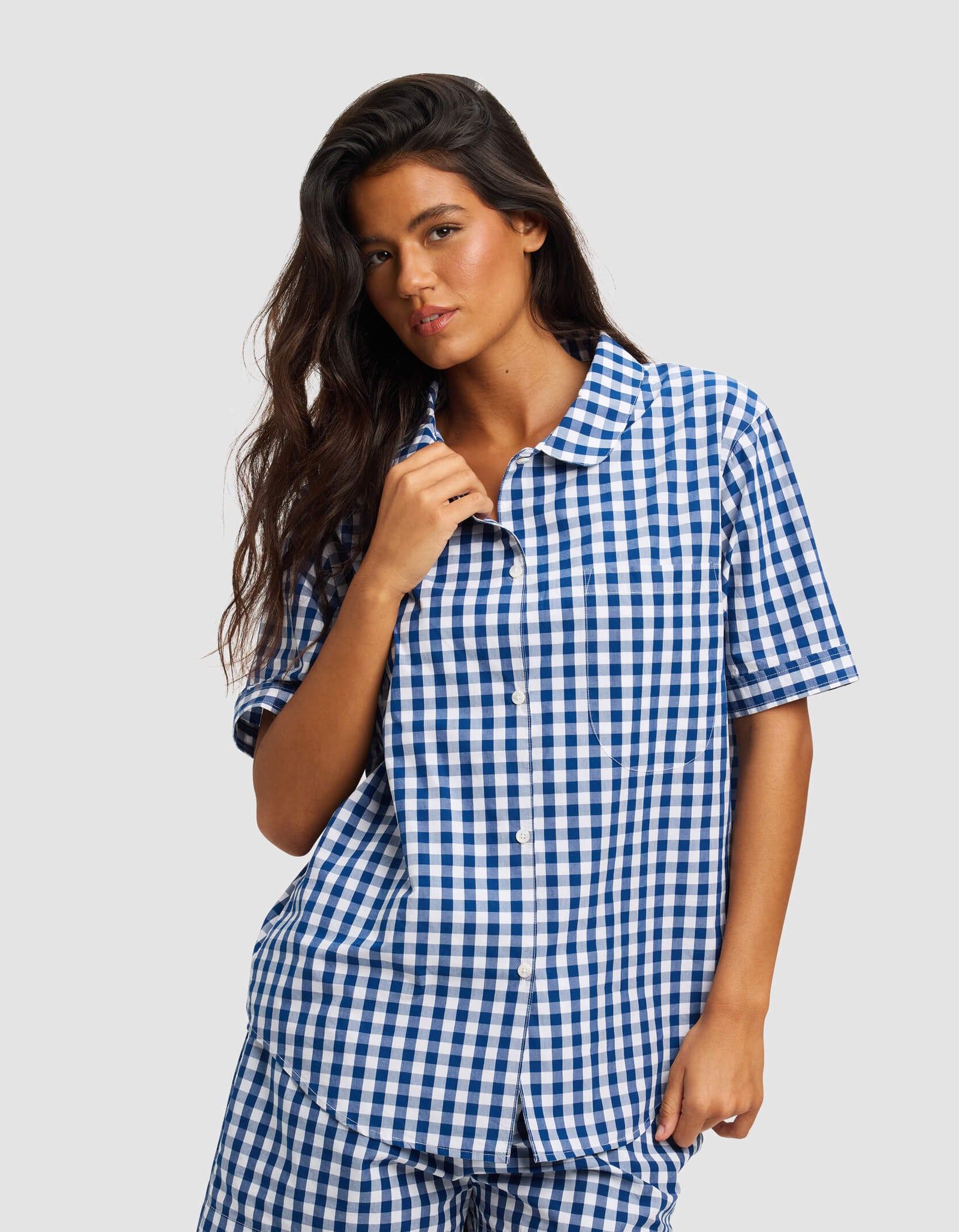 A woman with long dark hair wears the Cozy Earth Women's Sutton Short Sleeve Pajama Top, featuring a blue and white checkered pattern, standing against a plain light gray background.