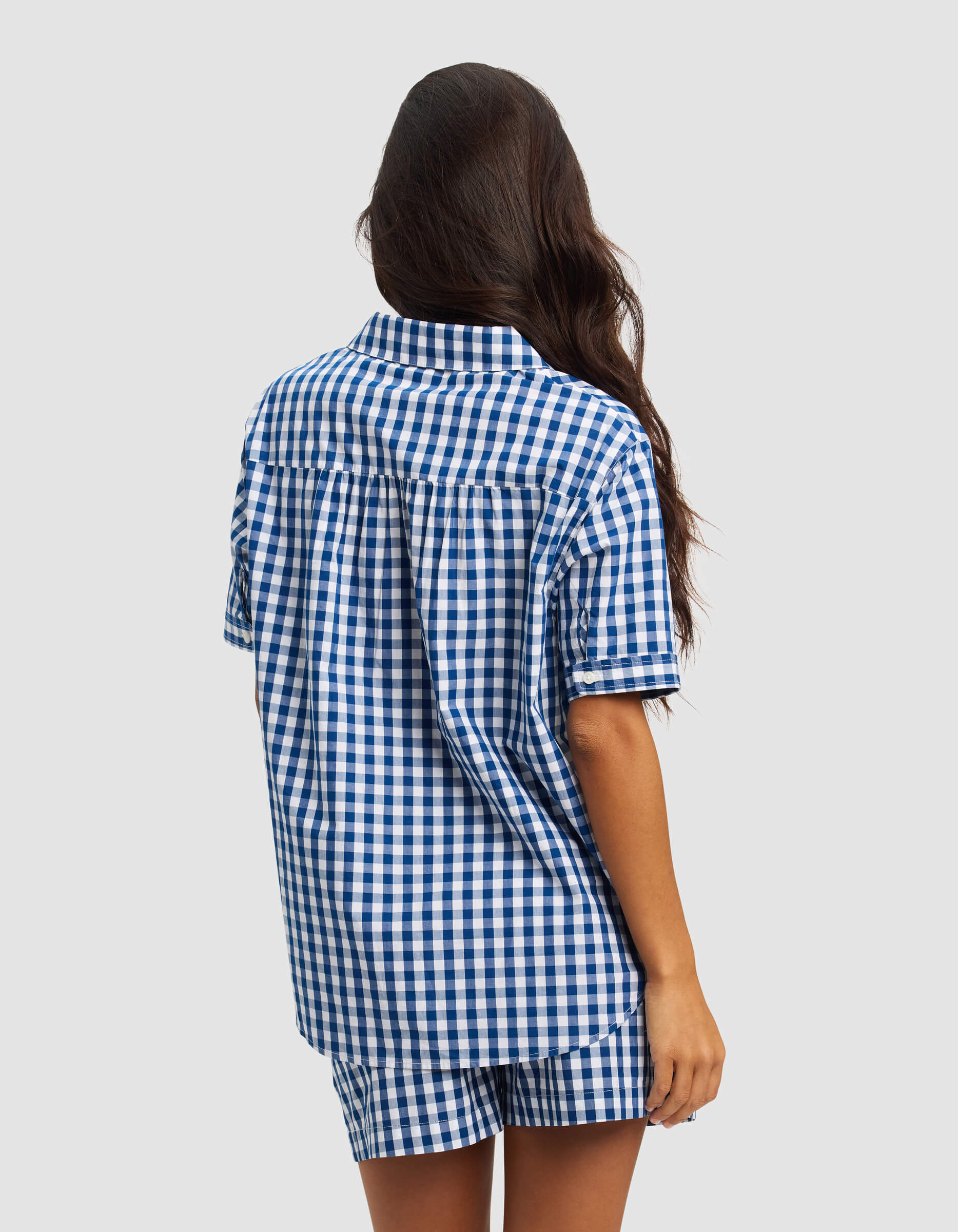 A woman with long, wavy hair faces away, wearing Cozy Earth's Women's Sutton Short Sleeve Pajama Top in a blue and white check pattern with matching shorts, set against a plain light background.|Color:Pacific Gingham