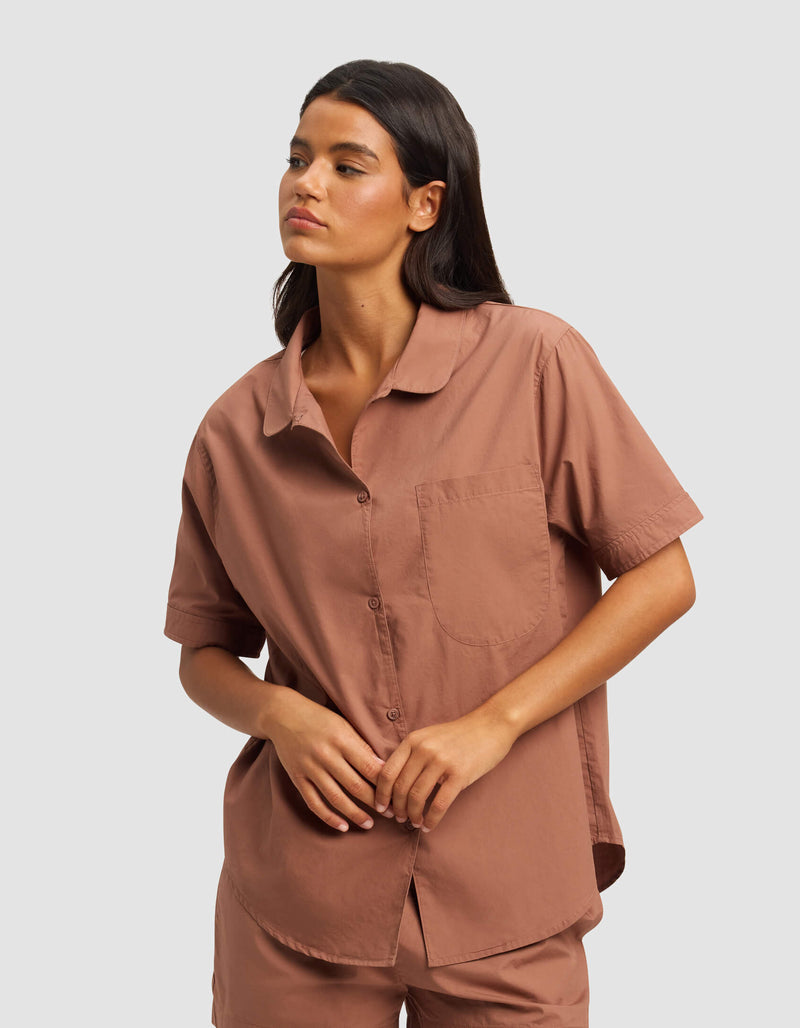 A woman with long dark hair wears the Cozy Earth Women's Sutton Short Sleeve Pajama Top and matching shorts in warm brown, standing with one hand resting on the other against a plain light gray background.