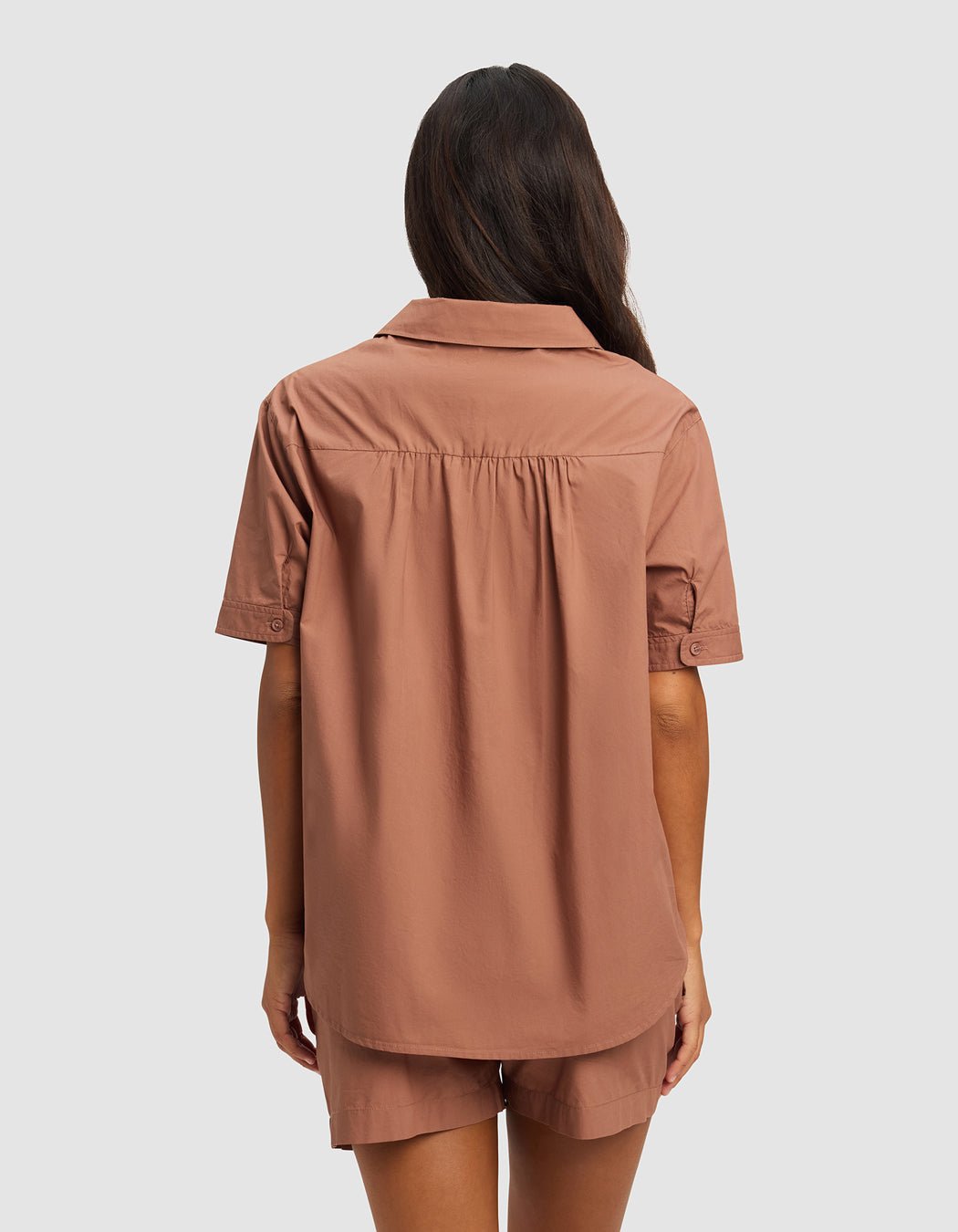 A woman with long dark hair is shown from the back, wearing the Cozy Earth Women's Sutton Short Sleeve Pajama Top in a light brown shade, paired with matching shorts, against a plain light background.
