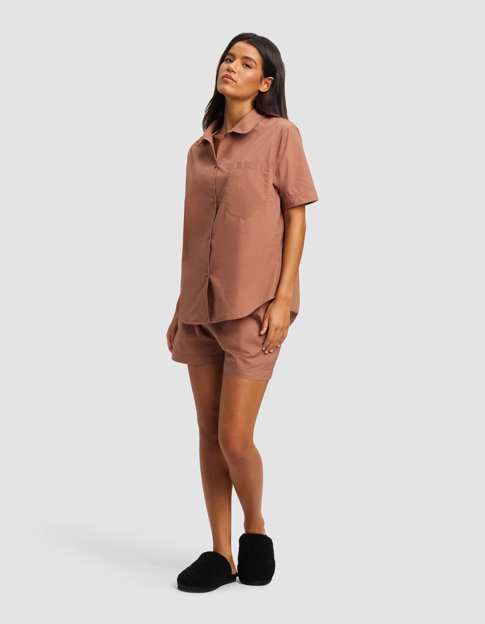 A woman stands against a plain background wearing the Cozy Earth Women's Sutton Short Sleeve Pajama Top in brown with matching shorts and black slippers.