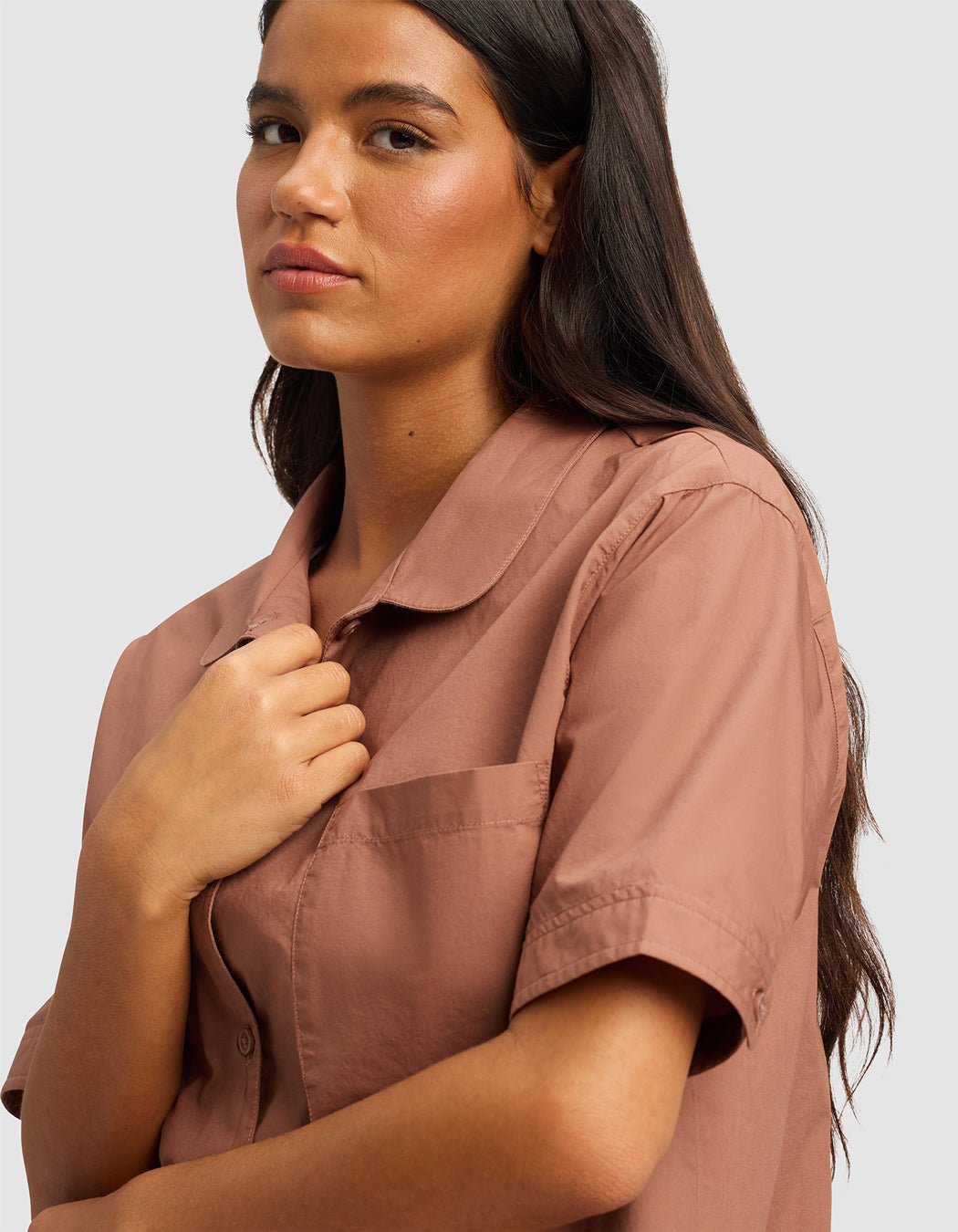 A woman with long dark hair stands against a light gray background, slightly looking to the side, wearing the Cozy Earth Women's Sutton Short Sleeve Pajama Top in light brown and touching the collar.