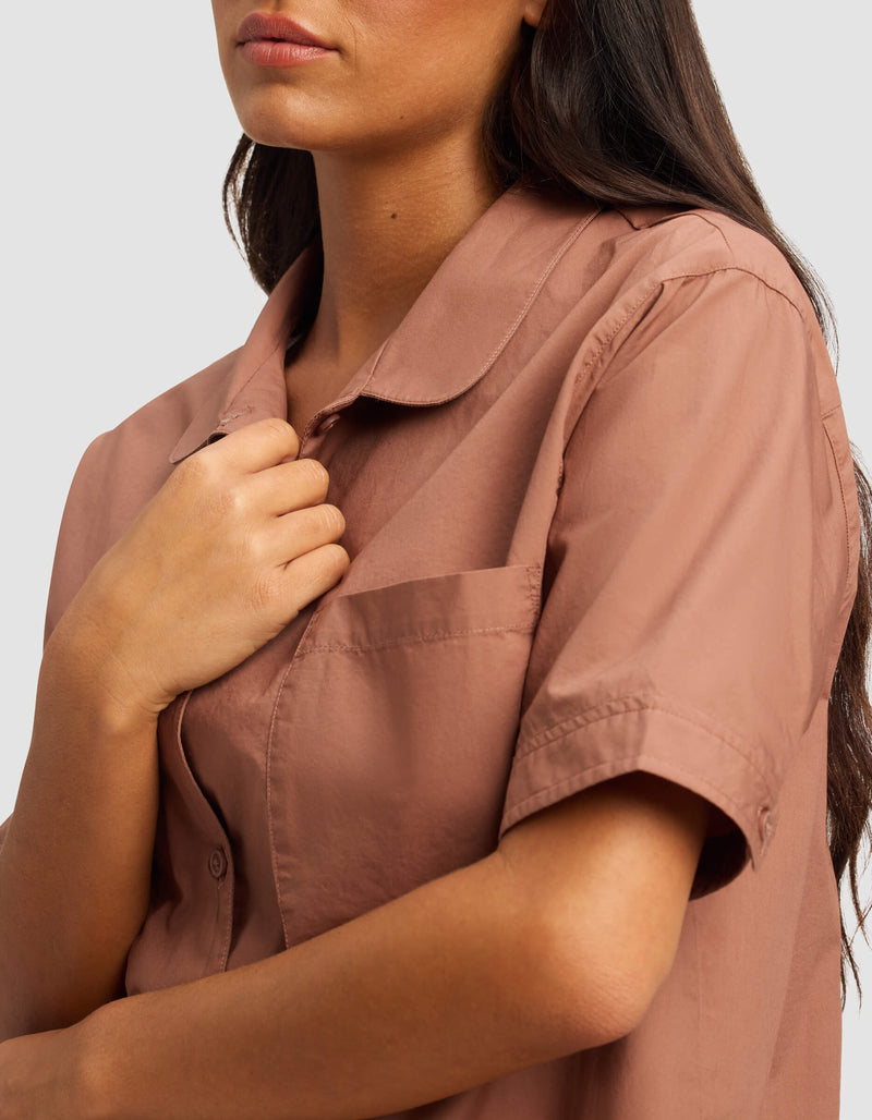 A woman with long brown hair wears the Cozy Earth Women's Sutton Short Sleeve Pajama Top in light brown, holding its collar with one hand. Her face is partially visible against a plain light gray background.