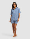 A woman stands barefoot against a plain light gray background wearing the Cozy Earth Women's Sutton Short Sleeve Pajama Set, featuring a matching blue and white checkered top and shorts.|Color:Pacific Gingham