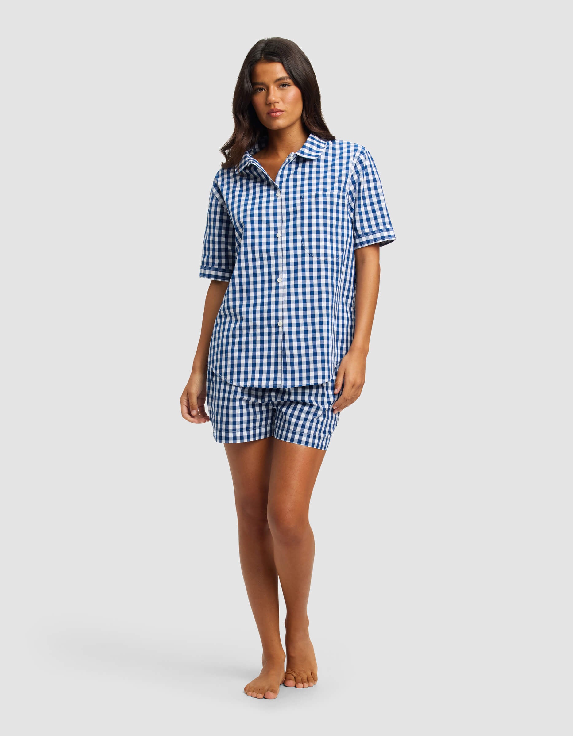 A woman stands barefoot against a light gray background wearing the Cozy Earth Women's Sutton Short Sleeve Pajama Set, featuring a matching blue and white gingham button-up shirt and shorts.|Color:Pacific Gingham
