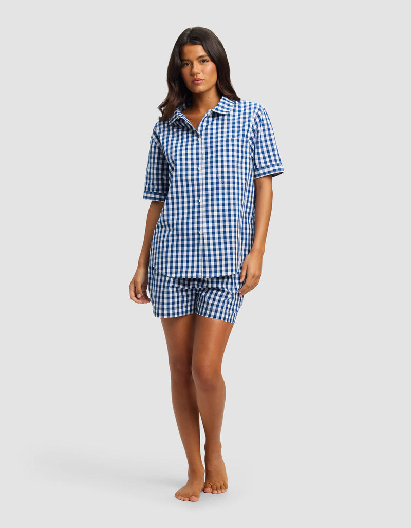 A woman stands barefoot against a light gray background wearing the Cozy Earth Women's Sutton Short Sleeve Pajama Set, featuring a matching blue and white gingham button-up shirt and shorts.