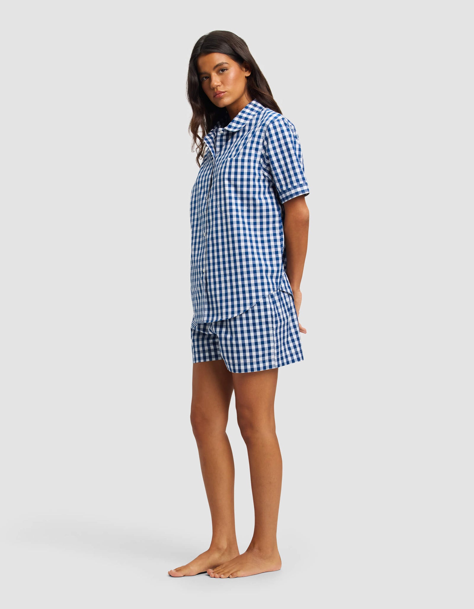 A woman stands barefoot against a light gray background, wearing Cozy Earth's Women's Sutton Short Sleeve Pajama Set, featuring a matching blue and white gingham button-up shirt and shorts.