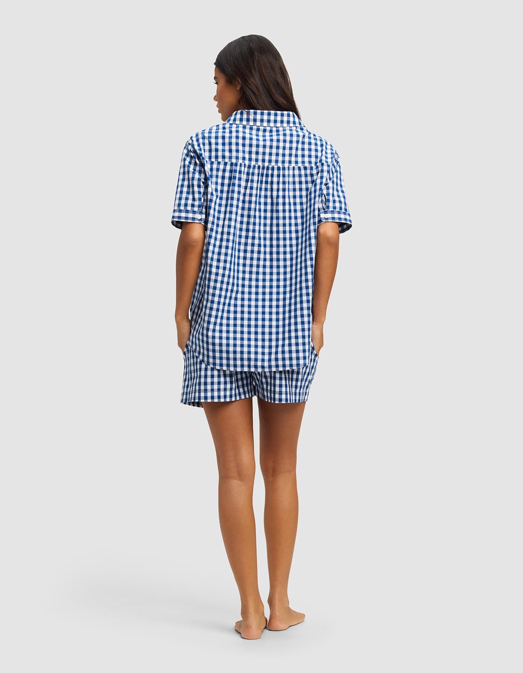 A woman stands barefoot facing away, wearing the Cozy Earth Women's Sutton Short Sleeve Pajama Set in a blue and white checkered pattern. The background is plain light gray.