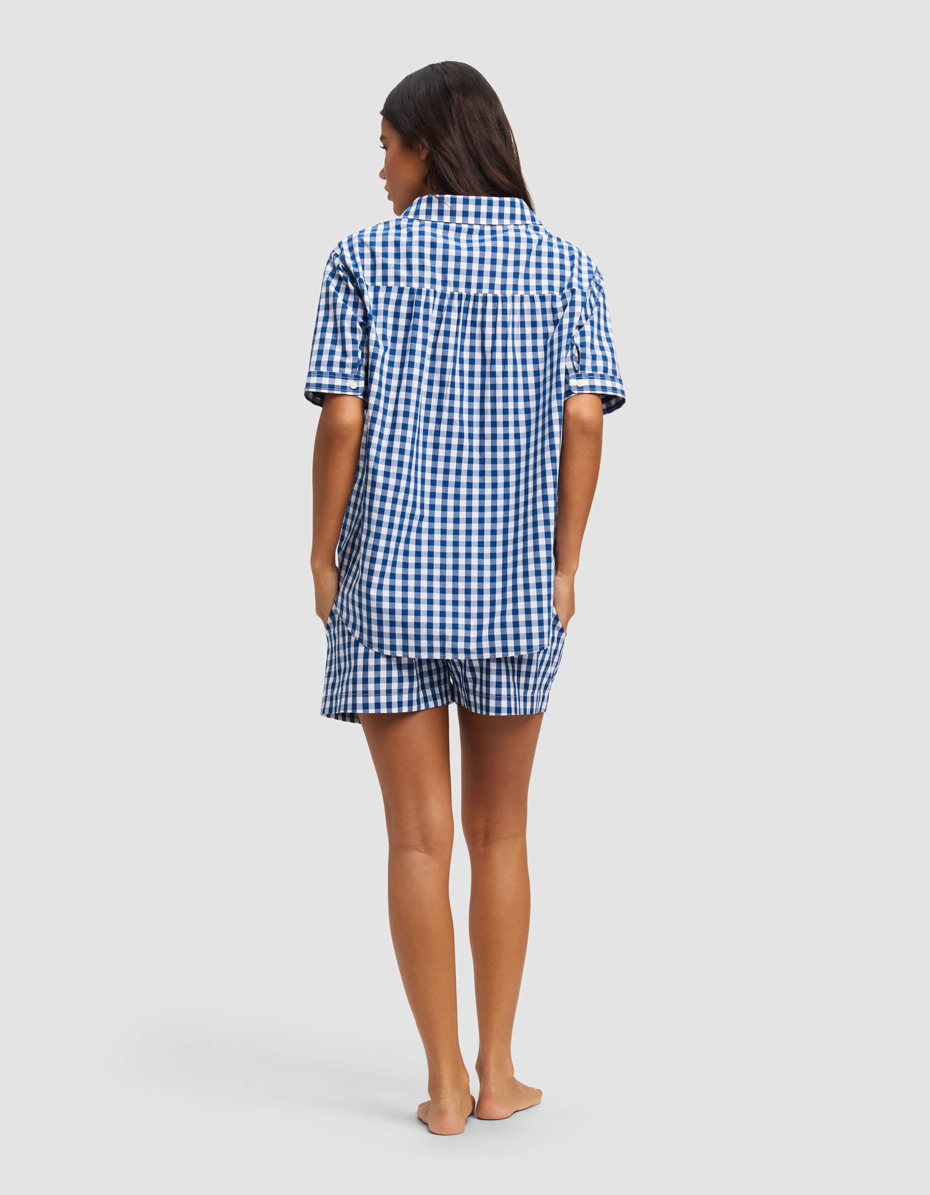 A woman stands barefoot, facing away, wearing the Cozy Earth Women's Sutton Short Sleeve Pajama Set—blue and white checkered top with matching shorts—against a plain, light-colored background.|Color:Pacific Gingham