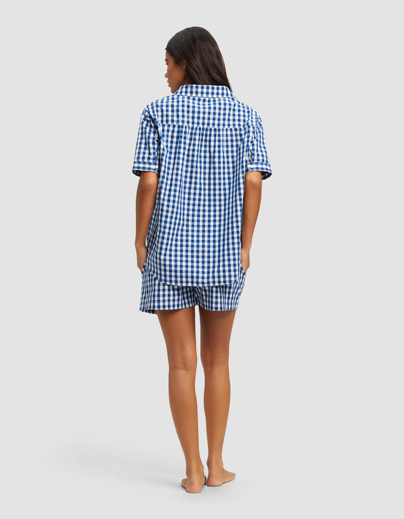 A woman stands barefoot, facing away, wearing the Cozy Earth Women's Sutton Short Sleeve Pajama Set—blue and white checkered top with matching shorts—against a plain, light-colored background.