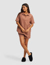 A woman smiles in the Cozy Earth Women's Sutton Short Sleeve Pajama Set—featuring a brown button-up top with matching shorts and black slippers—against a plain light gray background.|Color:Saddle
