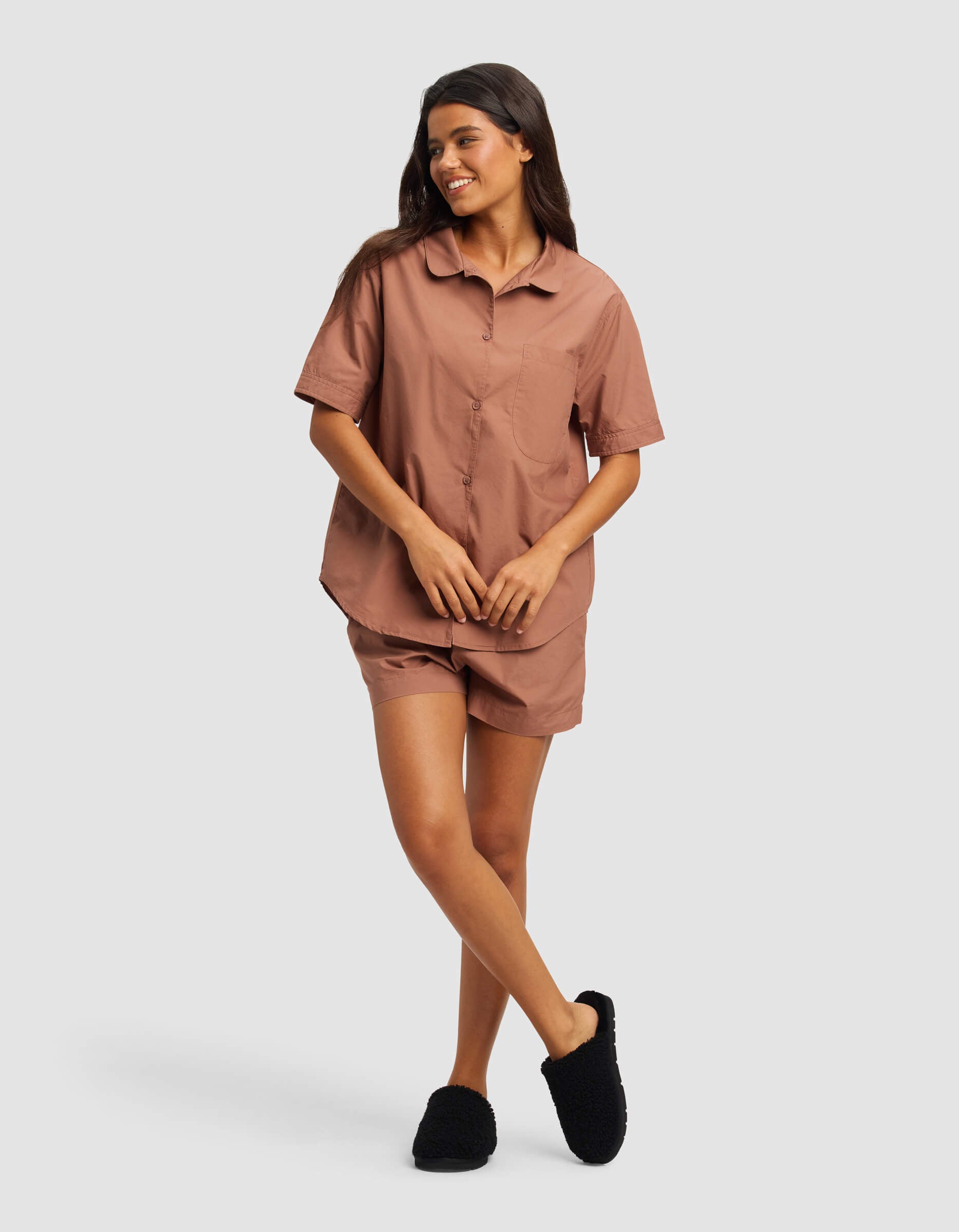 A woman smiles in the Cozy Earth Women's Sutton Short Sleeve Pajama Set—featuring a brown button-up top with matching shorts and black slippers—against a plain light gray background.|Color:Saddle
