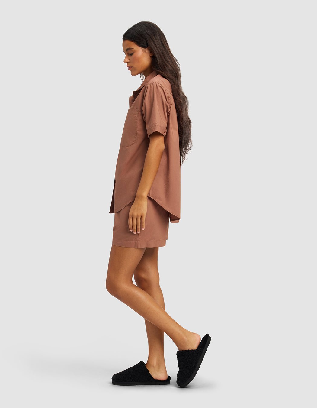 A woman with long dark hair models the Cozy Earth Women's Sutton Short Sleeve Pajama Set in brown, featuring a loose shirt and matching shorts, paired with black slippers, standing in profile against a light gray background.