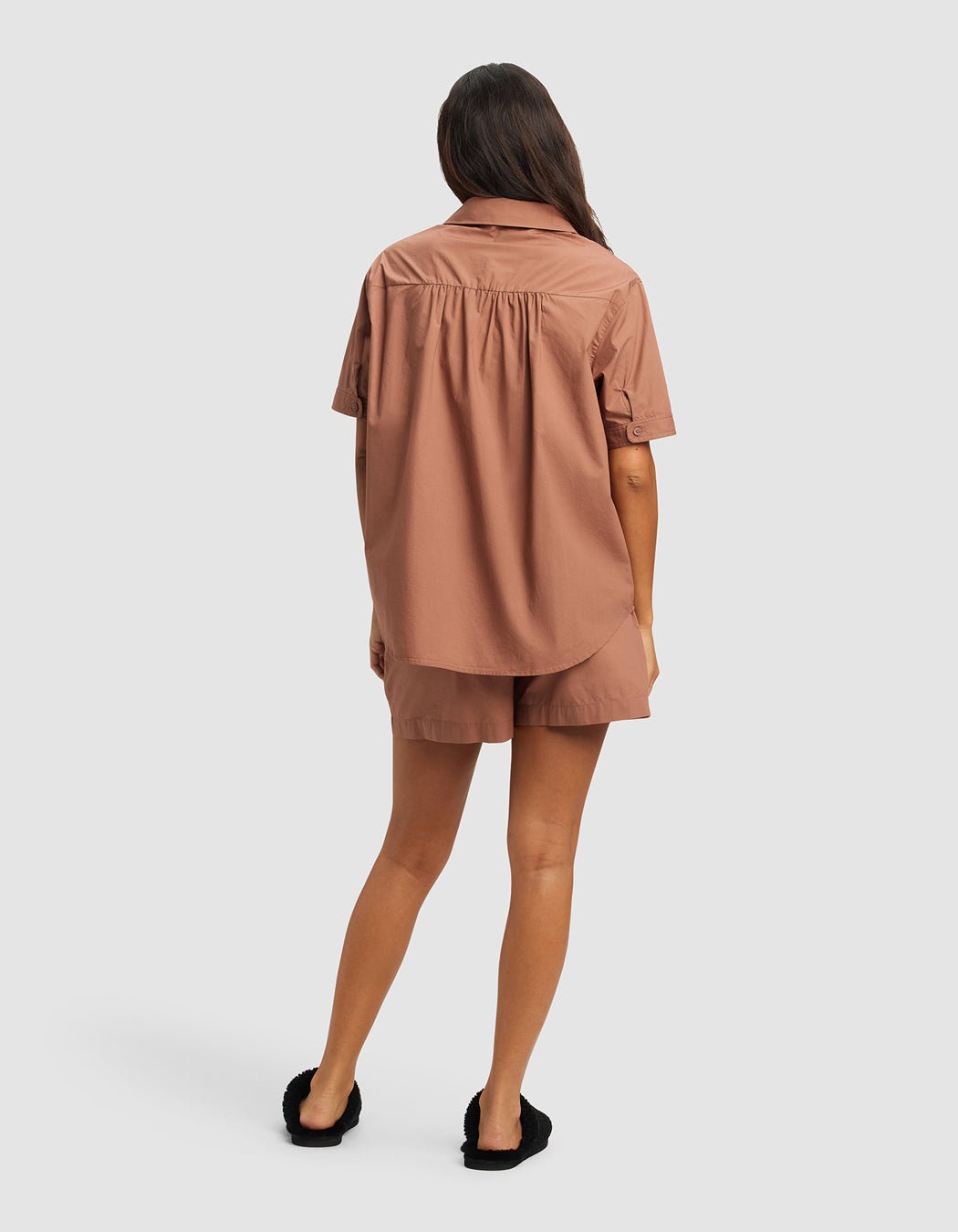 A woman with long brown hair wears the Cozy Earth Women's Sutton Short Sleeve Pajama Set in brown, featuring a relaxed-fit button-up shirt and matching shorts. She stands facing away in black slip-on shoes against a plain light gray background.