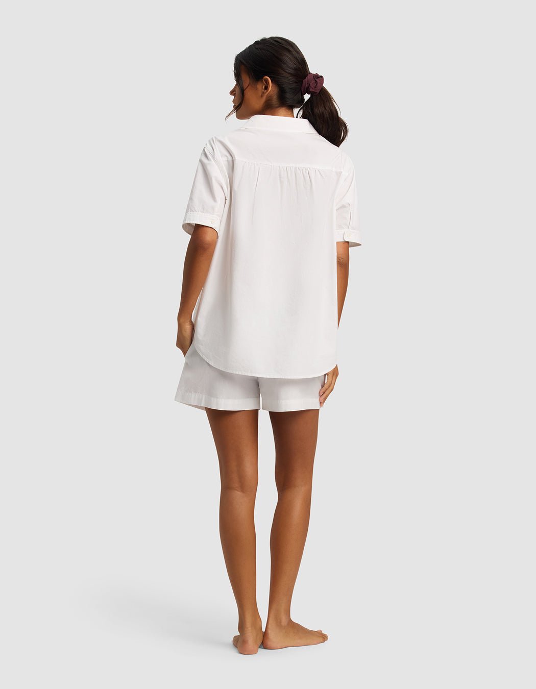 A woman with dark hair in a scrunchie stands barefoot, facing away, wearing the Cozy Earth Women's Sutton Short Sleeve Pajama Set—loose white shirt and matching shorts—against a plain light background.