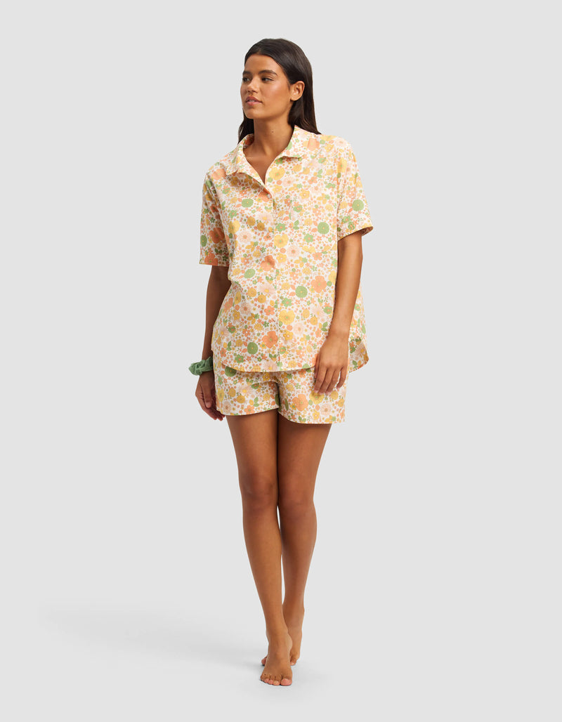 A woman stands barefoot in the Cozy Earth Women's Sutton Short Sleeve Pajama Set, featuring a matching button-up shirt and shorts with a pastel floral pattern, against a plain light gray background.
