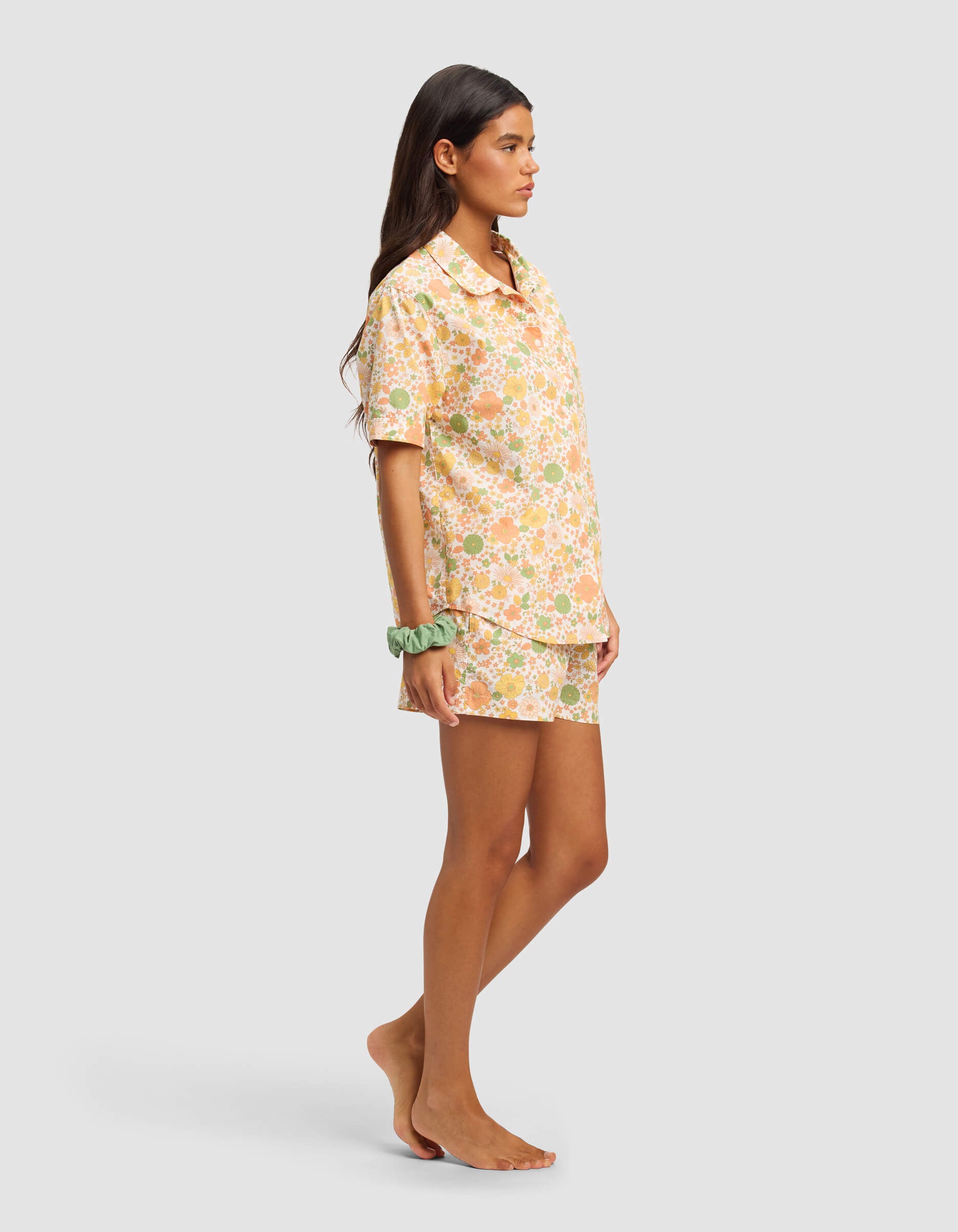 A woman with long brown hair stands barefoot, wearing the Cozy Earth Women's Sutton Short Sleeve Pajama Set with a peach and green floral pattern. She has a green scrunchie on her left wrist and faces left against a plain background.