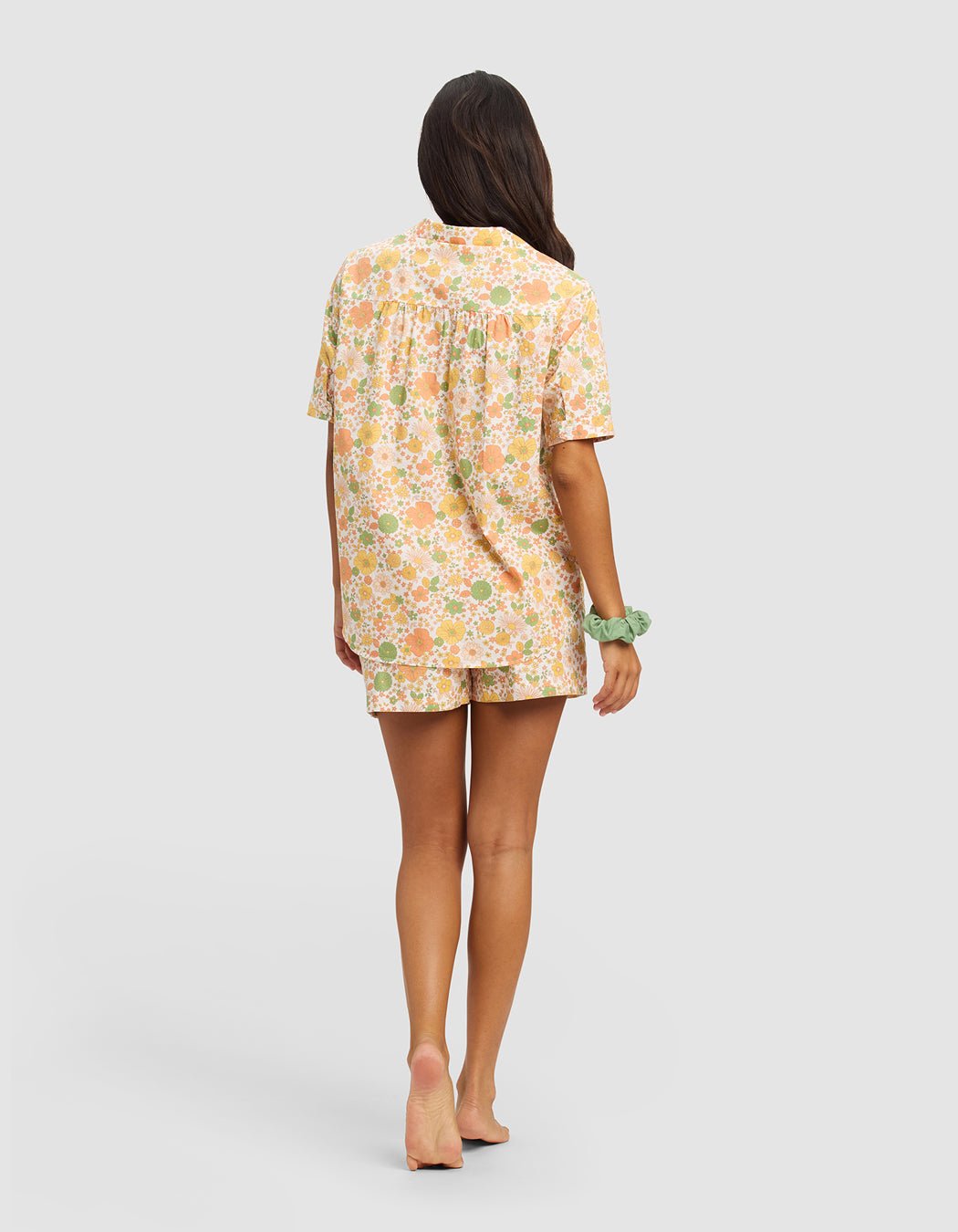 A woman with long dark hair stands barefoot, facing away, wearing the Women's Sutton Short Sleeve Pajama Set by Cozy Earth in a pastel floral print. She has a green scrunchie on one wrist against a plain light gray background.|Color:Wildflower