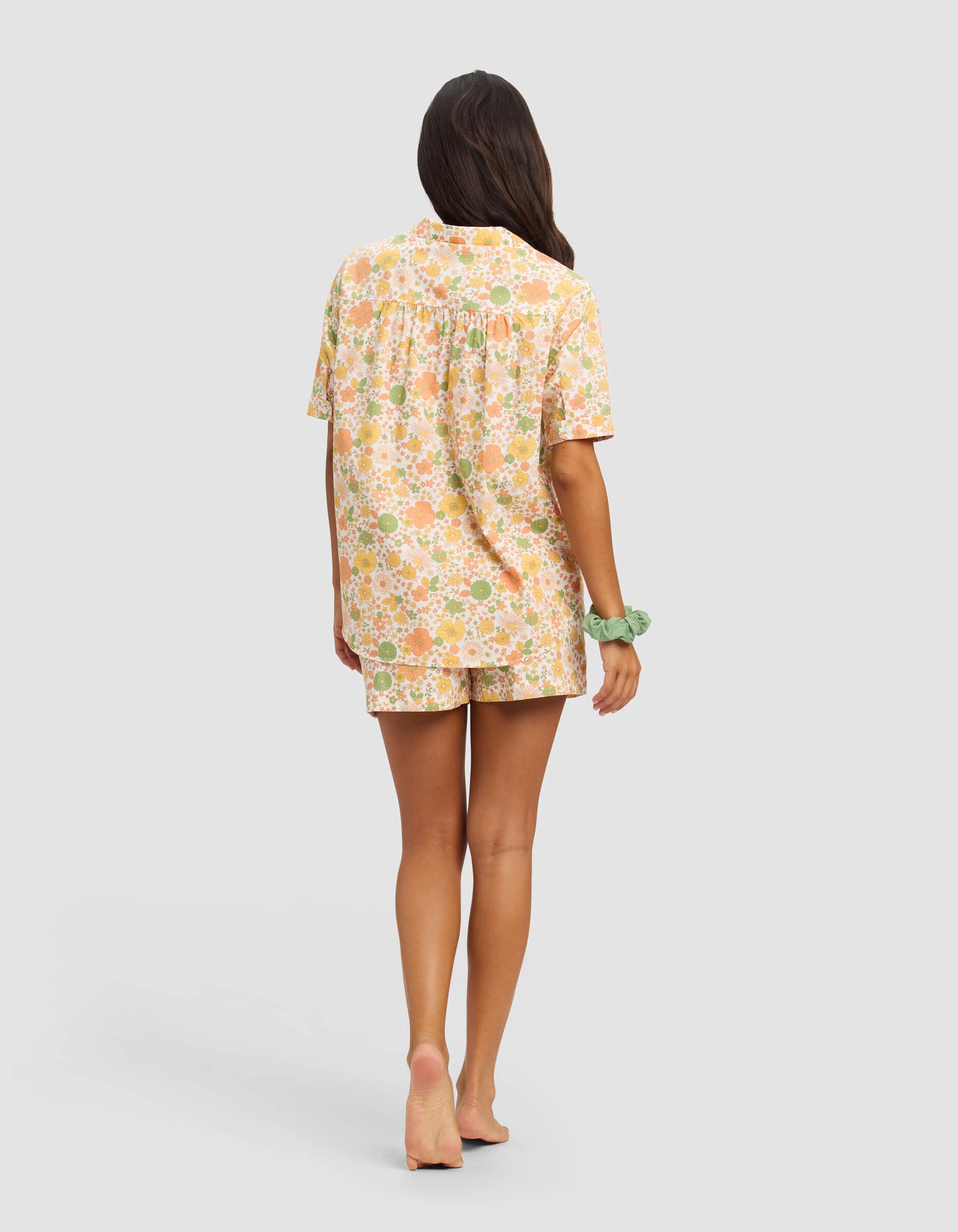 A woman with long brown hair, barefoot and facing away, wears the Cozy Earth Women's Sutton Short Sleeve Pajama Set in pastel orange, green, and white floral print, with a green scrunchie on her wrist against a plain background.