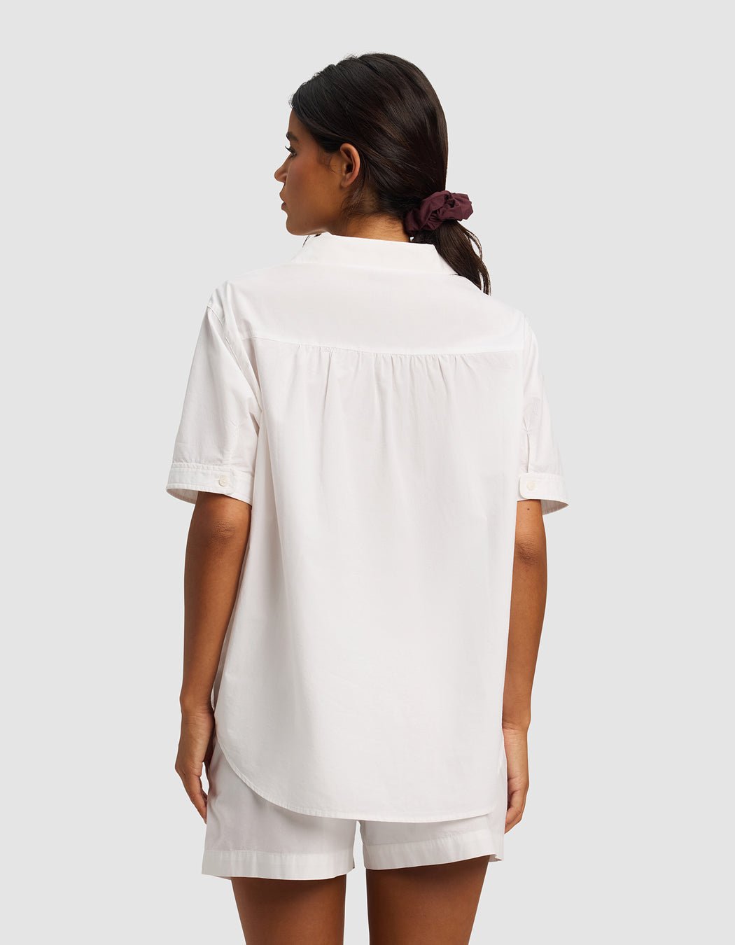 A woman, seen from behind, wears the Cozy Earth Women's Sutton Short Sleeve Pajama Top in white with matching shorts. Her hair is styled in a low ponytail with a dark scrunchie against a plain light gray background.