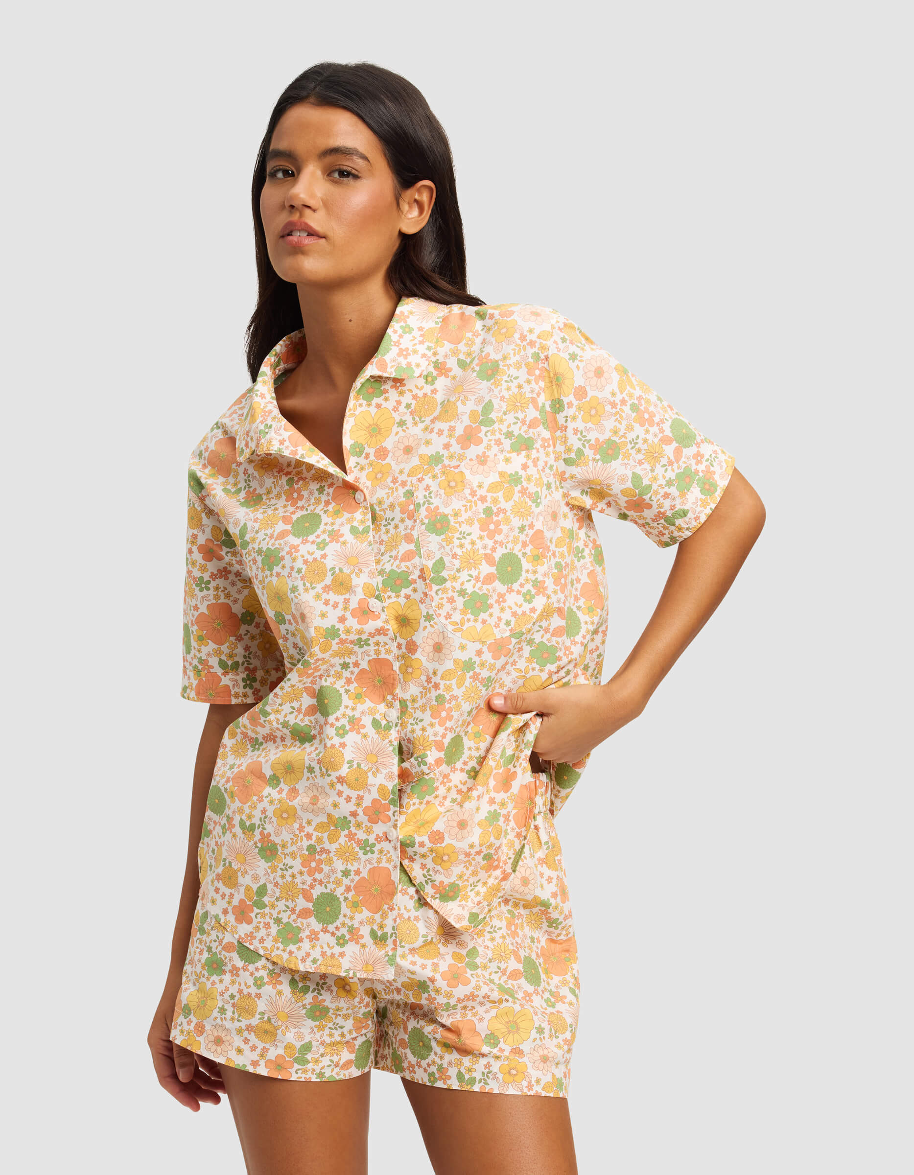A woman poses in the Cozy Earth Women's Sutton Short Sleeve Pajama Top with matching shorts, featuring a pastel orange, green, and white floral pattern. The background is light gray.