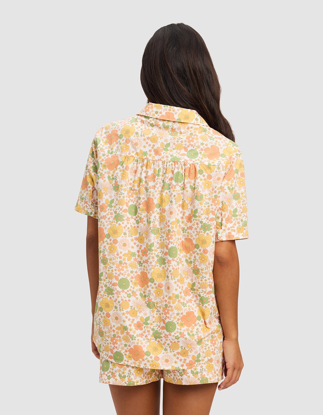 A woman with long dark hair stands facing away, wearing the Cozy Earth Women's Sutton Short Sleeve Pajama Top featuring a colorful yellow, orange, and green floral print on a light background.|Color:Wildflower